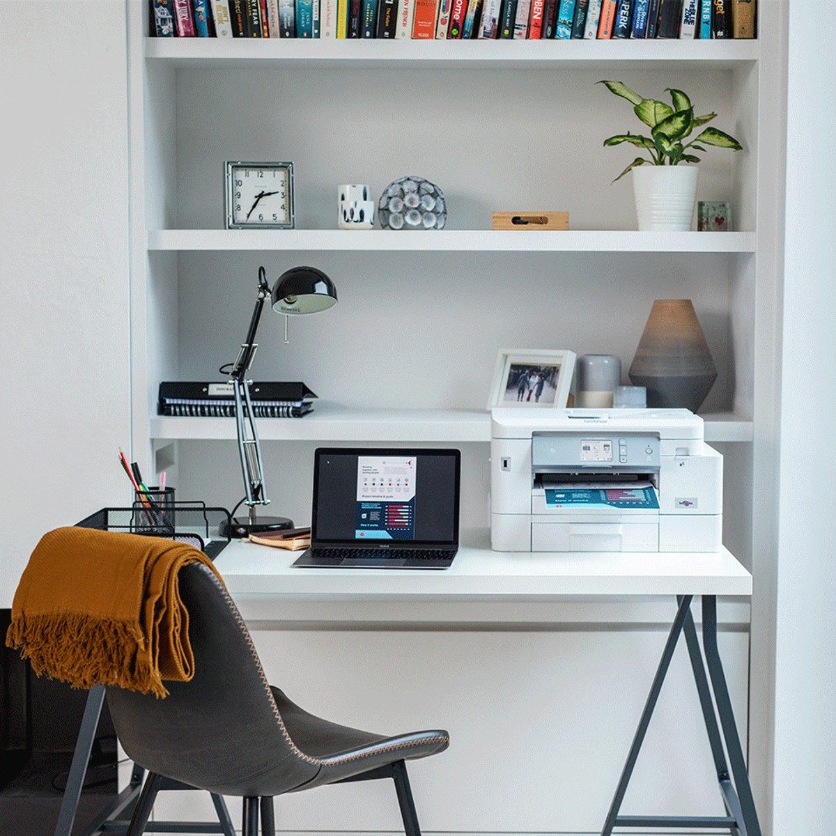 Escritorio con impresora, portátil y decoración. Estanterías al fondo.