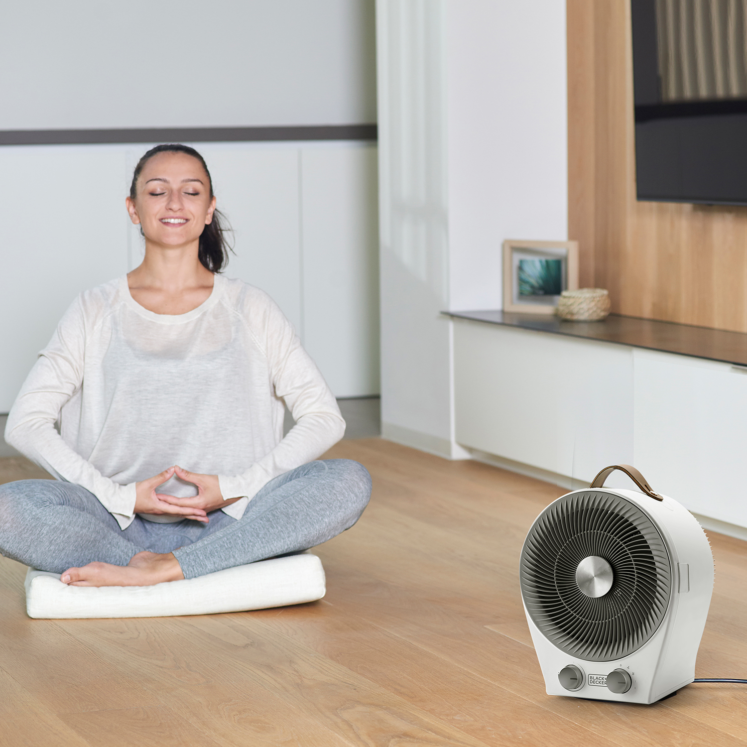 Kobieta medytuje na poduszce z białym wentylatorem na pierwszym planie. Drewniana podłoga.