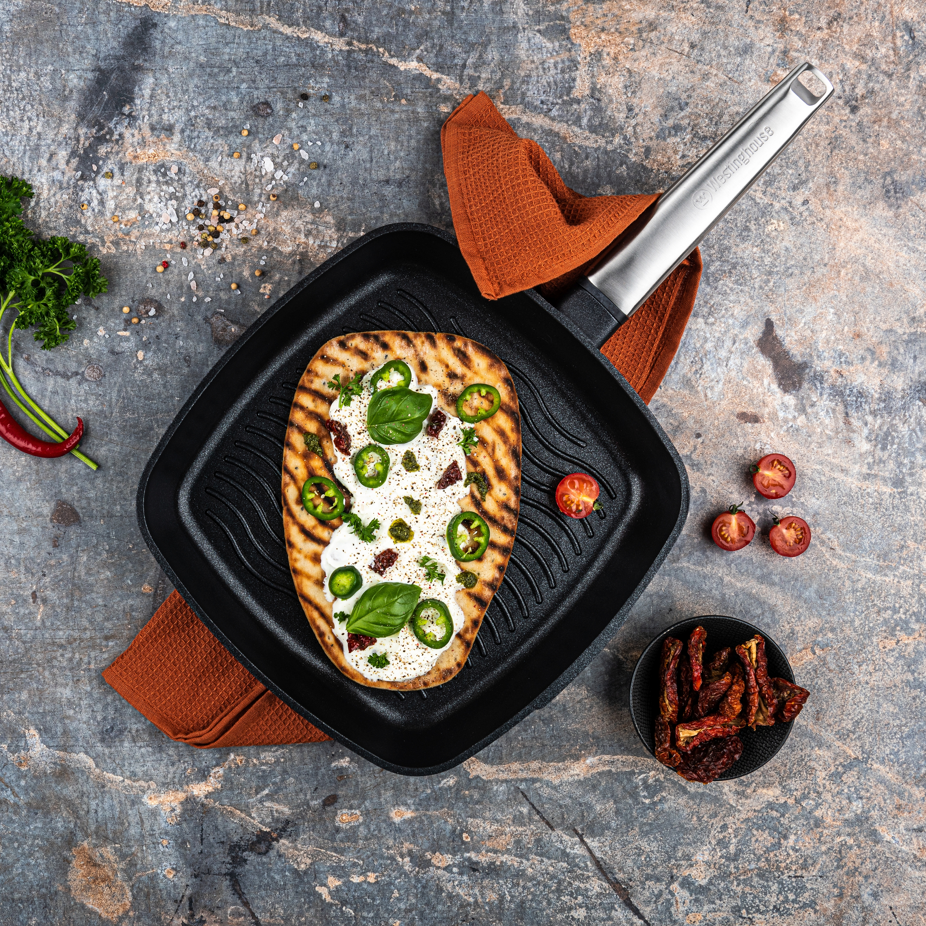 Ein Fladenbrot mit Belägen auf einer Grillpfanne, mit Tomaten, Kräutern und Zutaten drumherum.