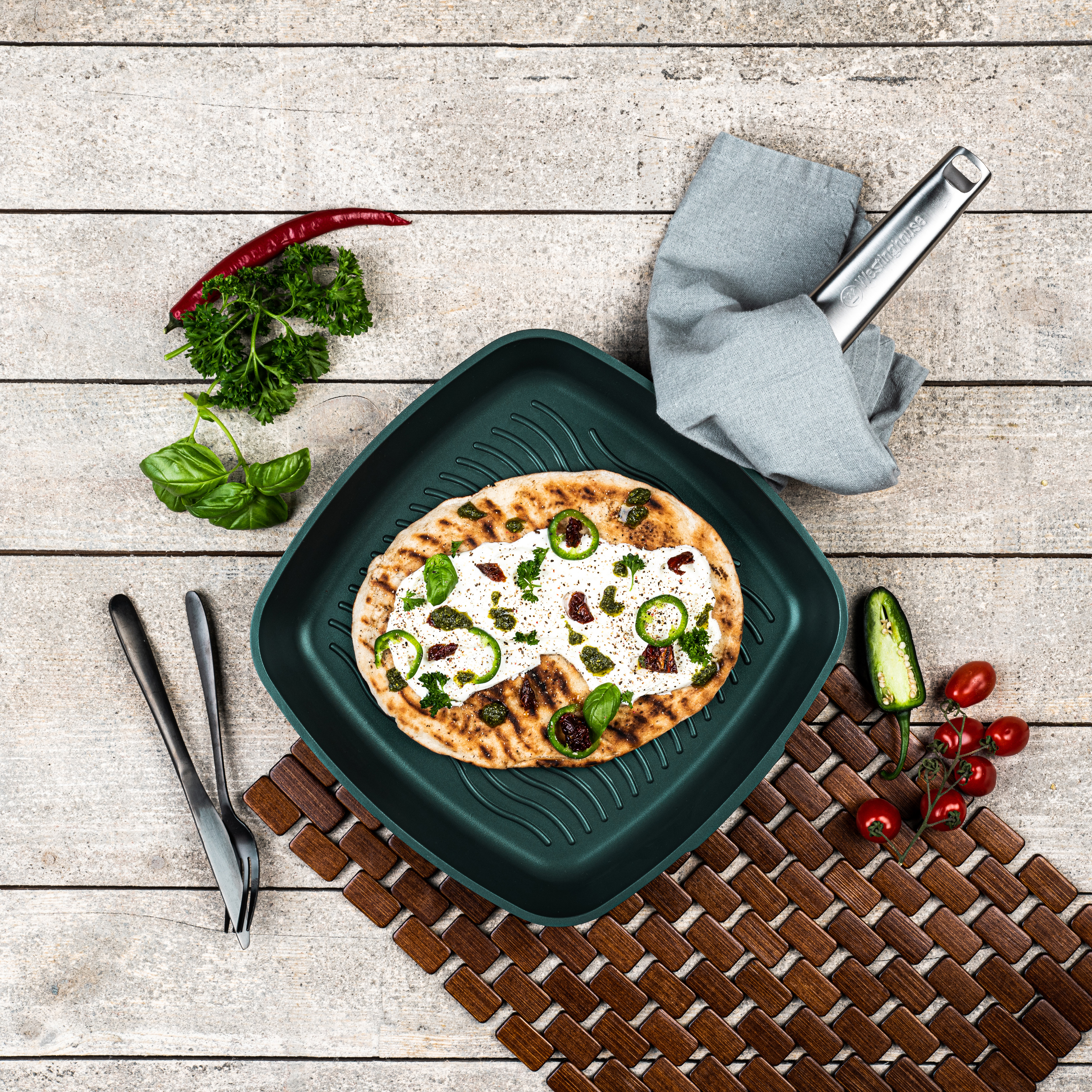 Pizza mit Belag auf einer grünen Grillpfanne, mit Utensilien und Zutaten.