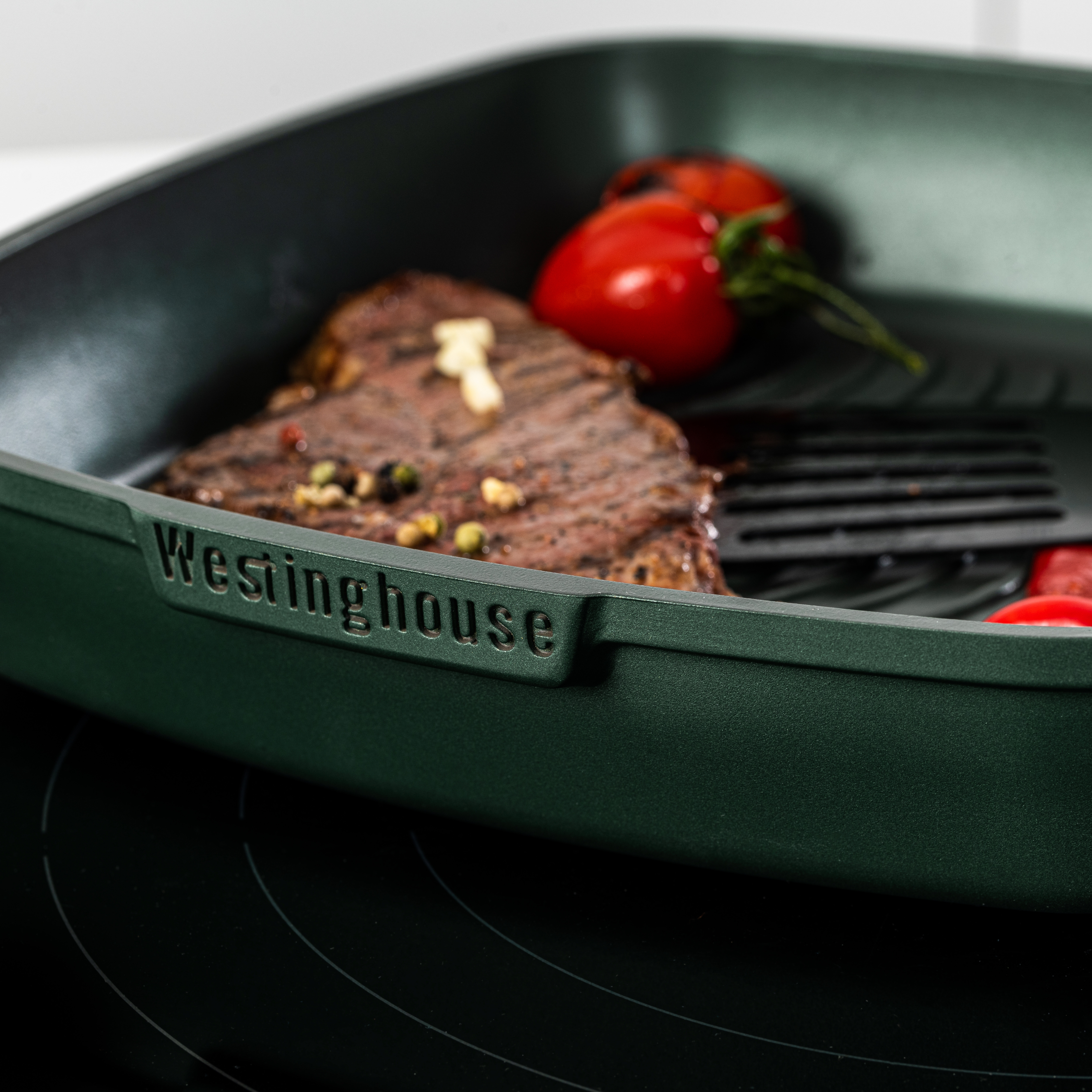 Grüne Grillpfanne mit Steak und Tomatenscheiben. Die Pfanne steht auf einem schwarzen Herd.