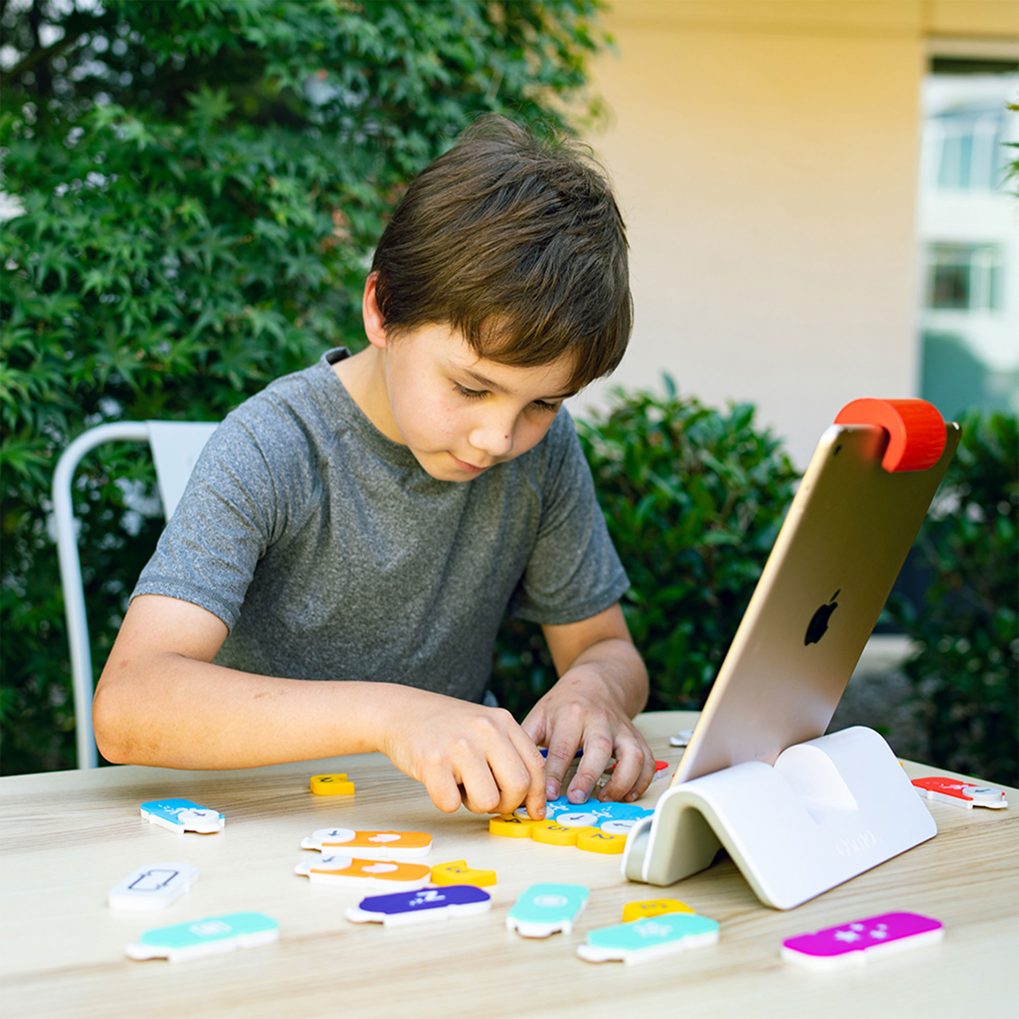 Un niño usa una tableta con bloques coloridos en una mesa al aire libre en un jardín.