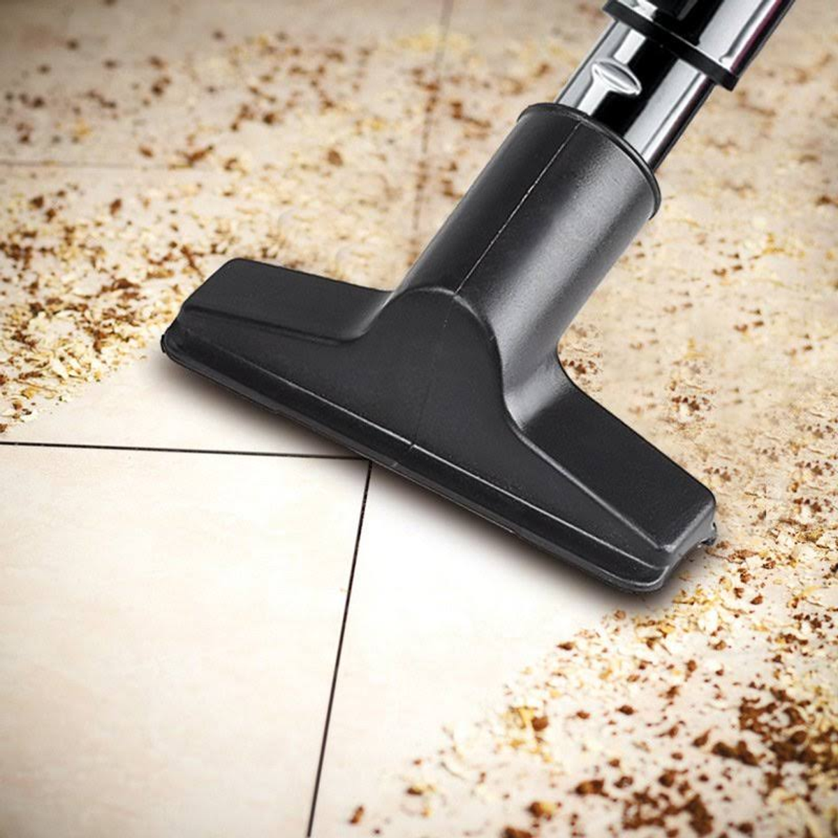 Zbliżenie głowicy odkurzacza czyszczącej beżową podłogę z płytek. Brązowe resztki rozsiane wokół.