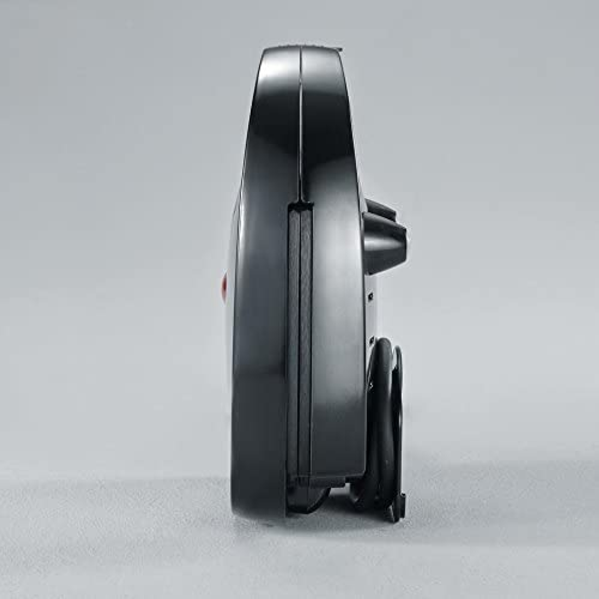 Black waffle maker, side view. Rounded design. Power cord visible. Gray background.