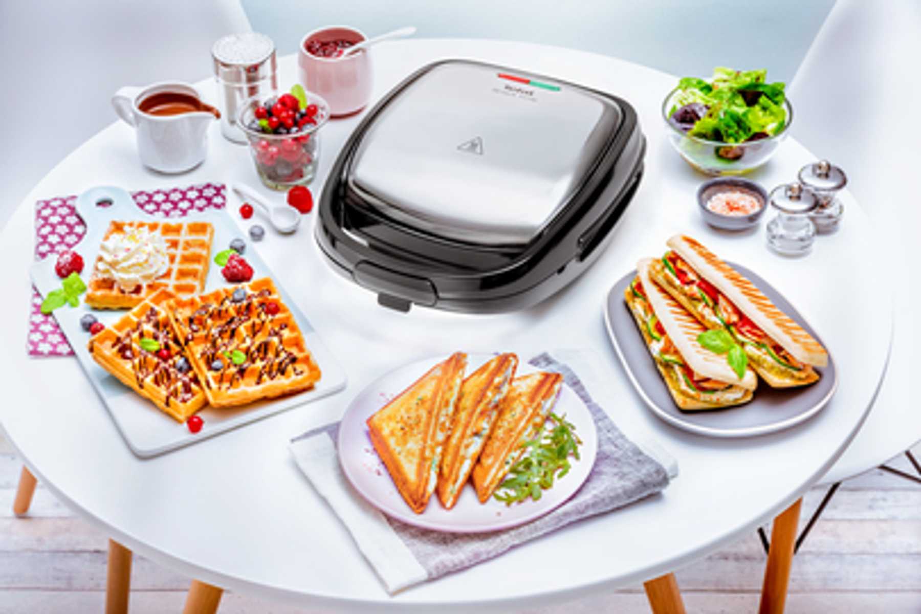 Tisch mit Waffeleisen, Waffeln, Sandwiches, Salat und Gewürzen. Weißer Tisch.