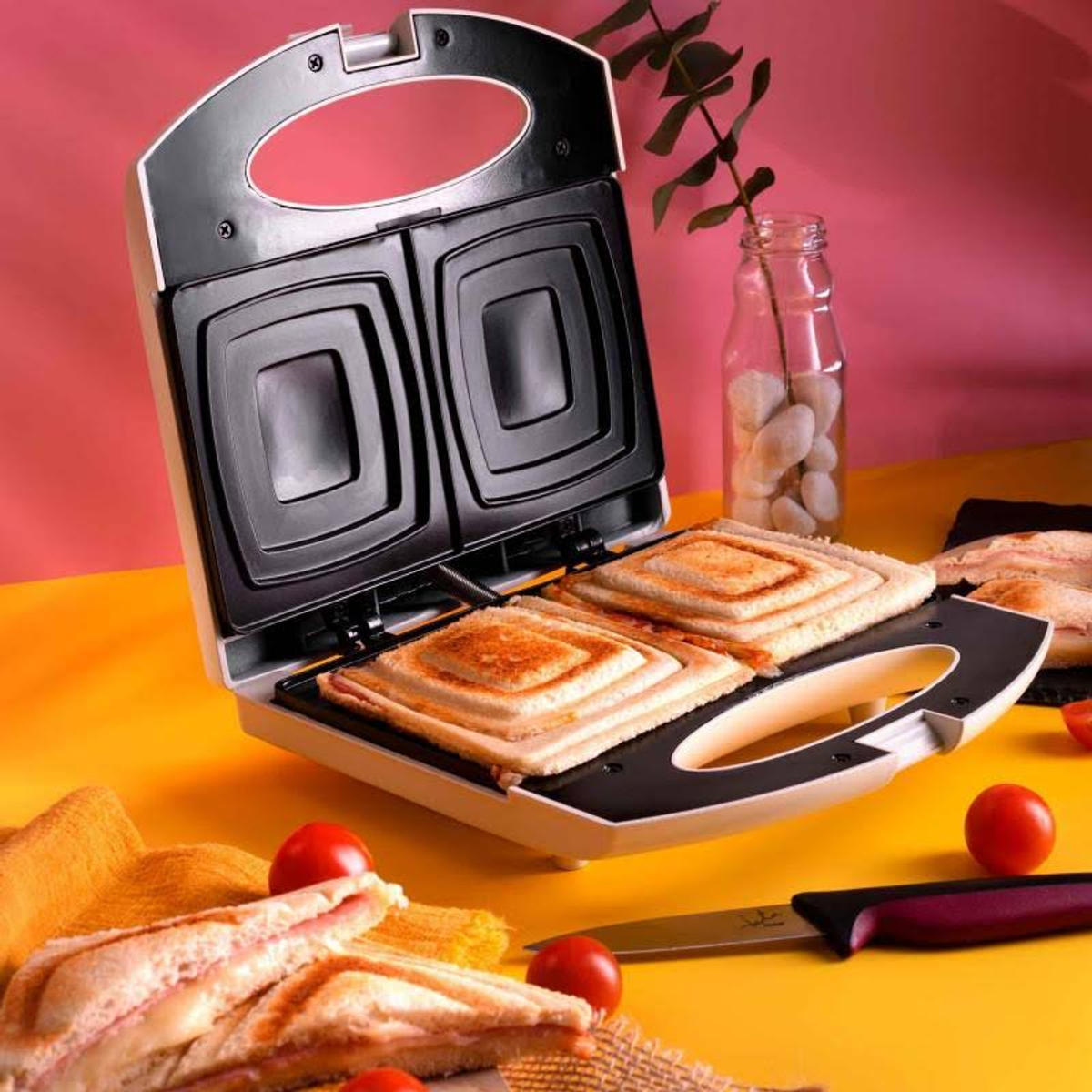 Panini maker aperto con due panini tostati. Inoltre, pomodori e un coltello sono sul tavolo.