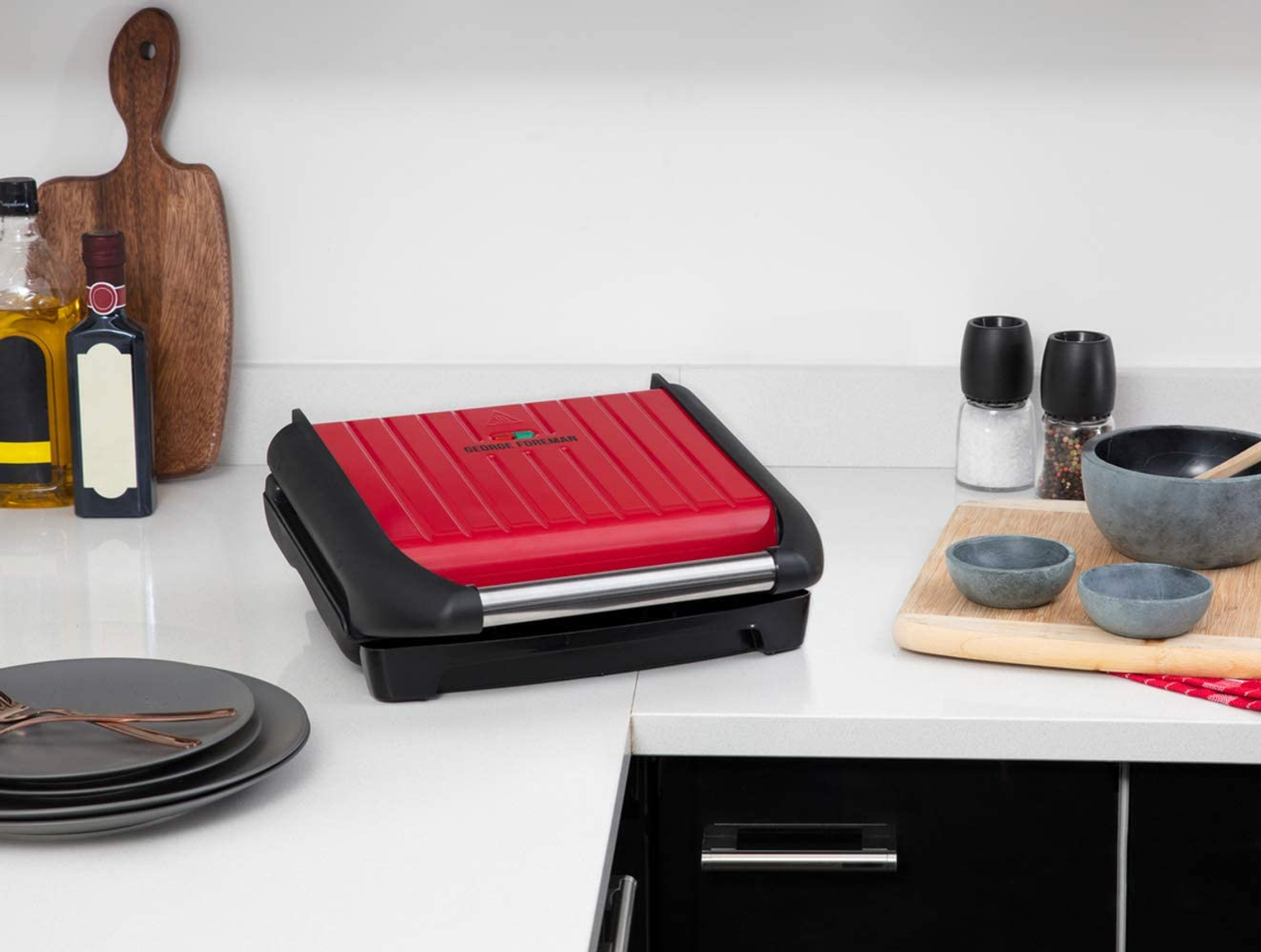 Griglia George Foreman rossa e nera su un piano di lavoro bianco, oggetti da cucina sullo sfondo.