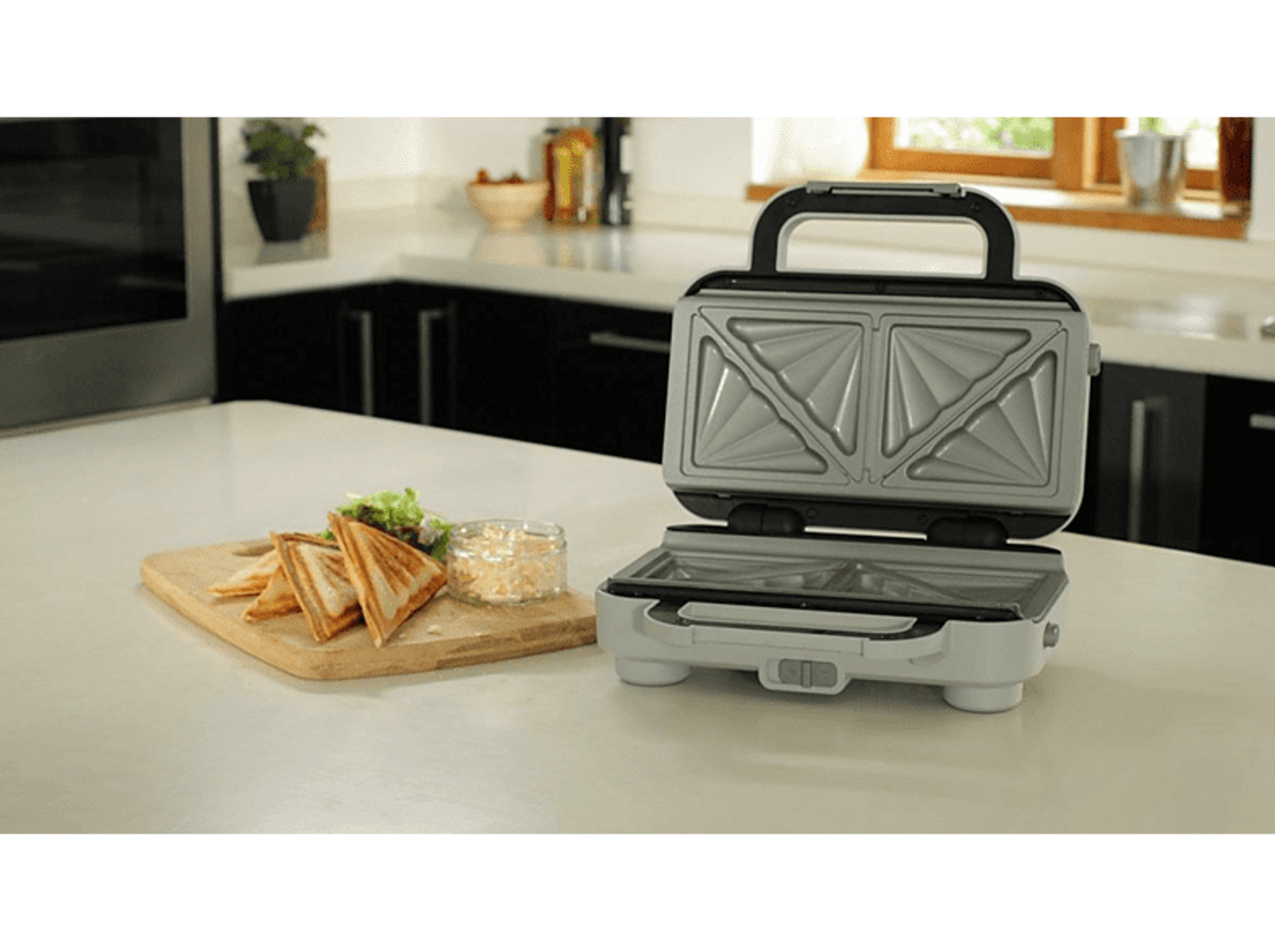 Ein offener Sandwichmaker mit Sandwiches auf einem Holzbrett. Küchenhintergrund.