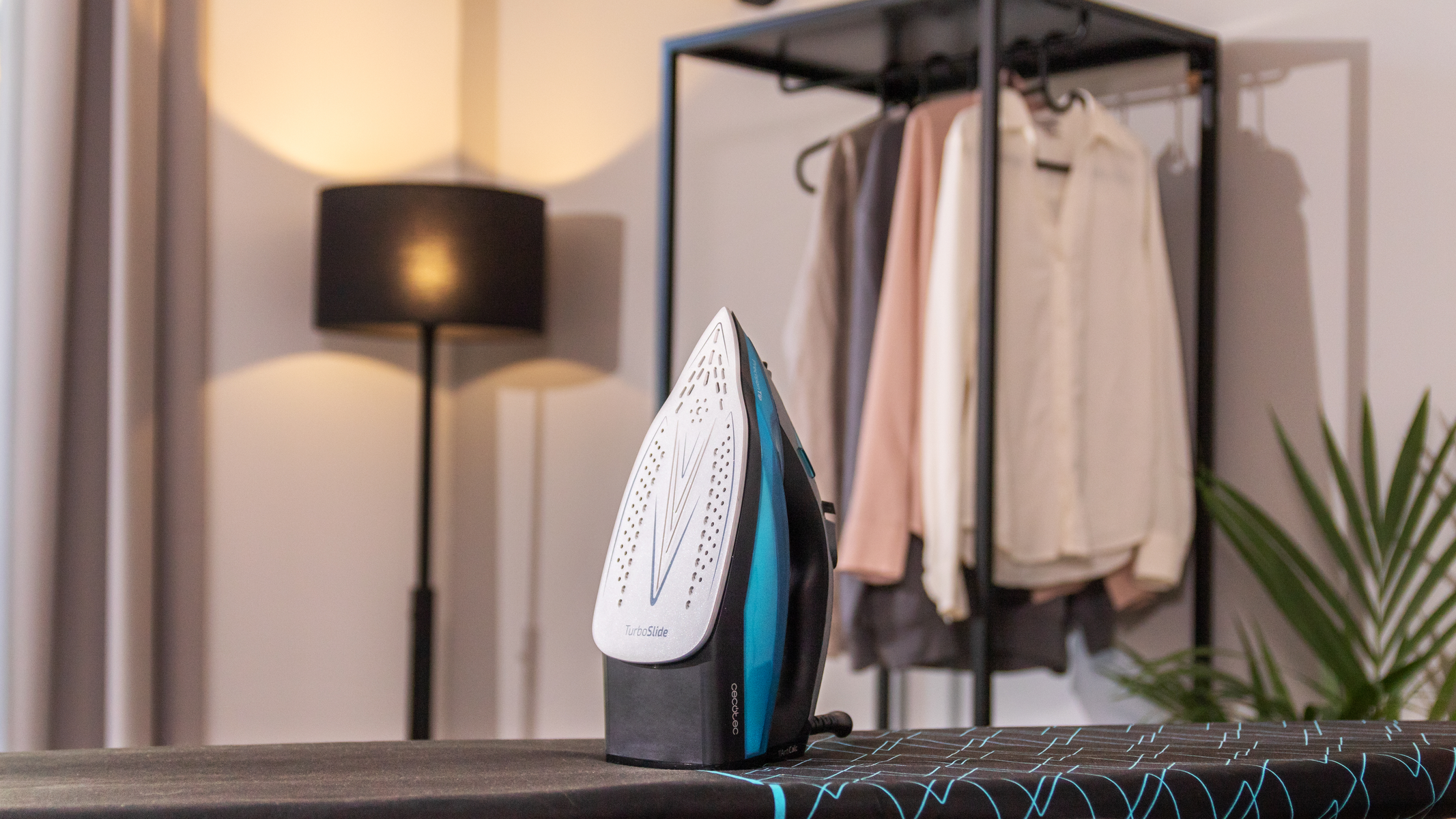 Żelazko na desce do prasowania z ubraniami w tle.
