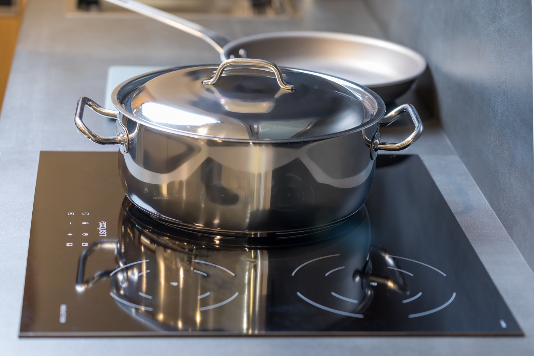 Olla de acero inoxidable con tapa en vitrocerámica negra de inducción, con una sartén al fondo.