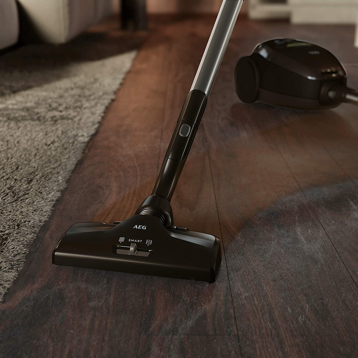 Czarny odkurzacz z długim uchwytem, na drewnianej podłodze. Osobna jednostka w tle.