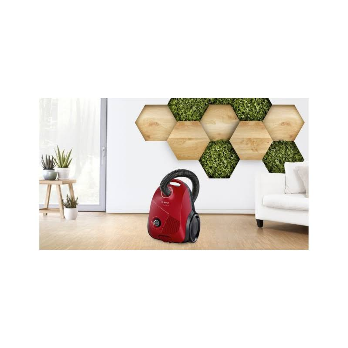 Czerwony odkurzacz stoi na jasnej podłodze. Dekoracyjna ściana z drewna i roślin za nim.