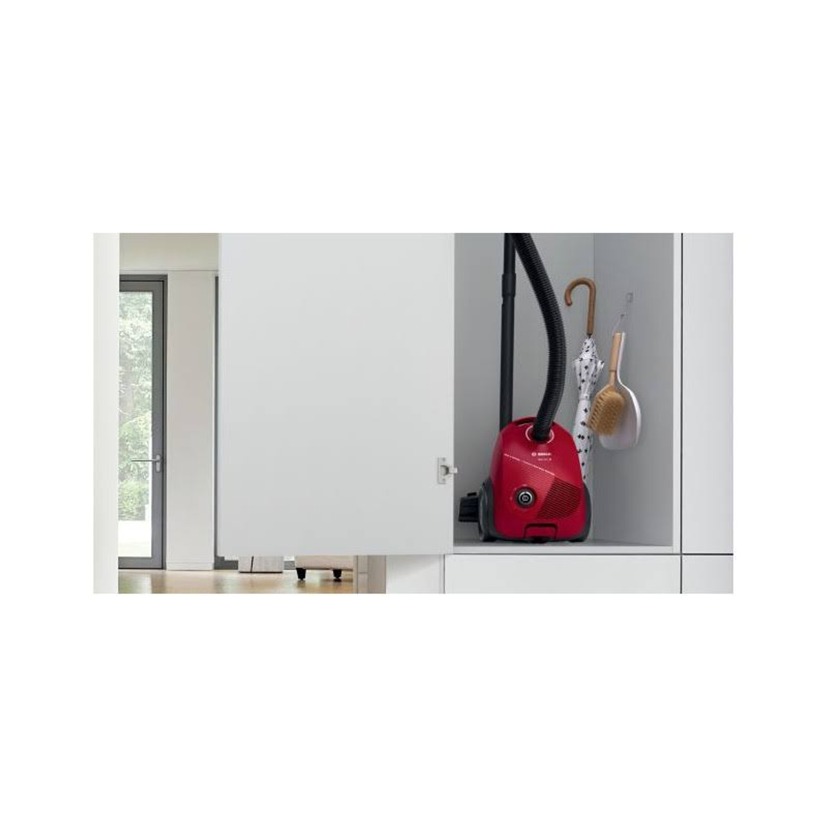 Czerwony odkurzacz w białej szafce. Drzwi są uchylone. Wiszą parasol i szczotka.