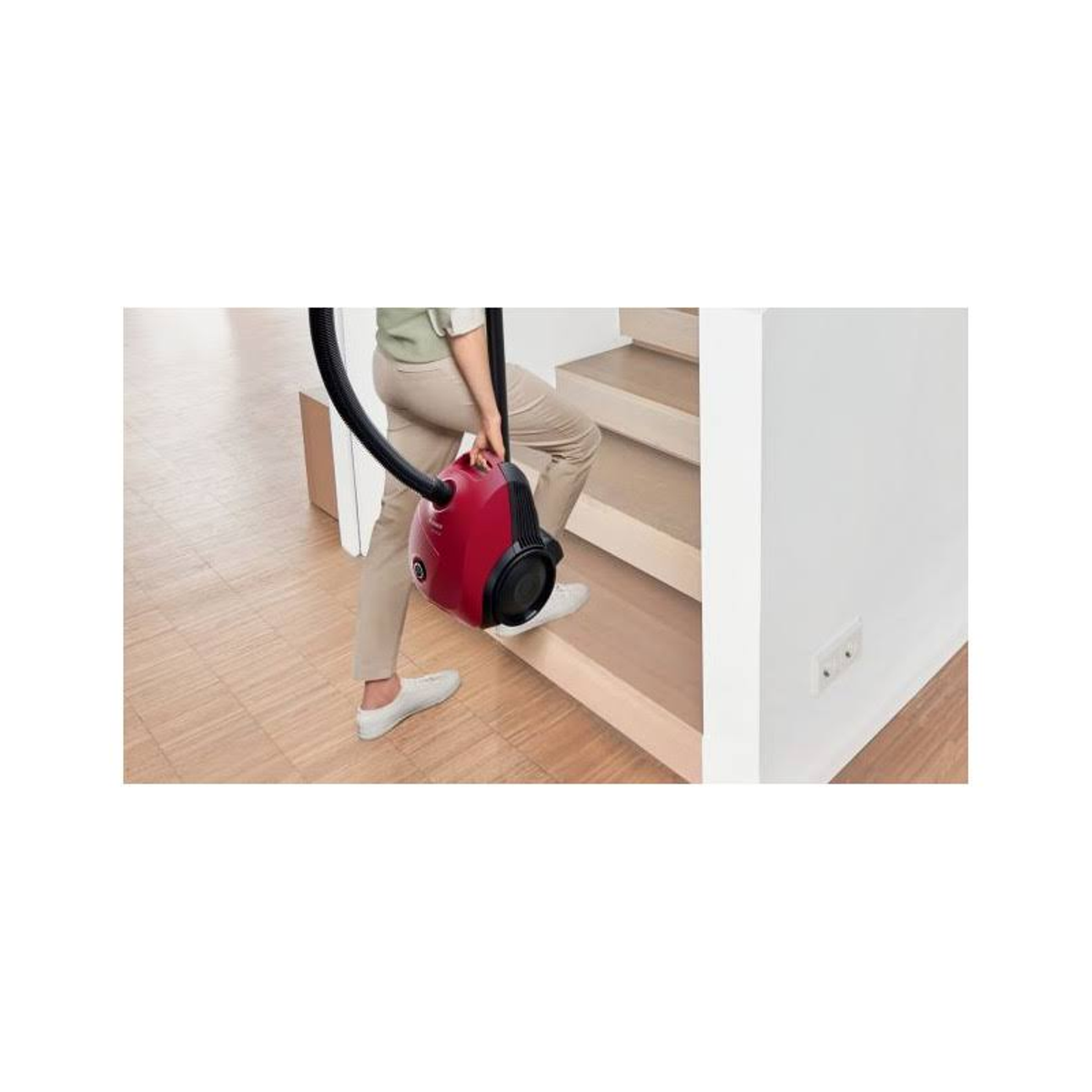 Osoba niosąca czerwony odkurzacz po drewnianych schodach, w beżowych spodniach i białych butach.