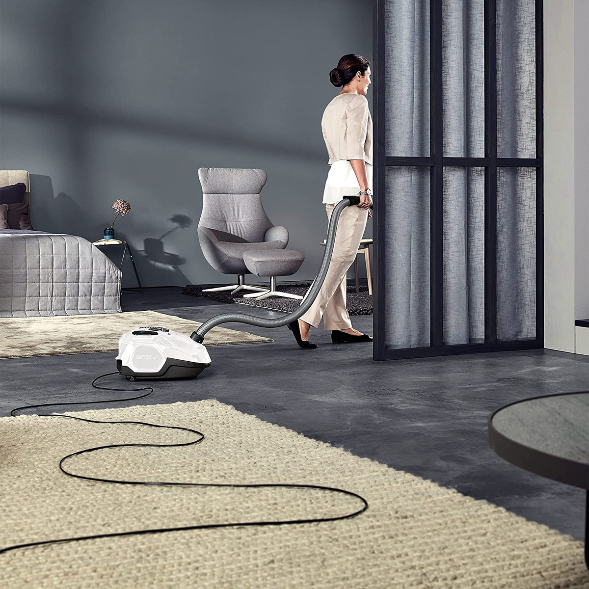 Kobieta odkurzająca pokój białym odkurzaczem i szarym wężem, na szarej podłodze.
