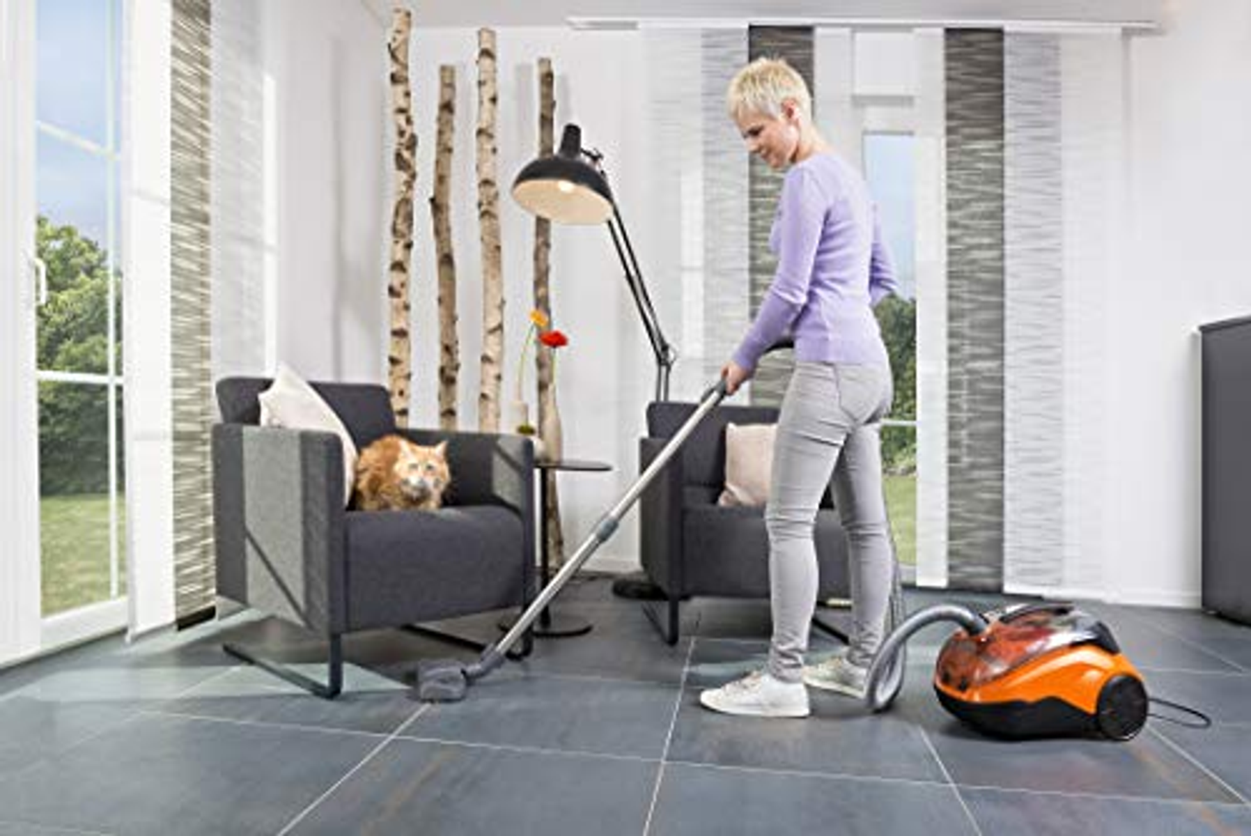 Kobieta odkurza pokój. Pomarańczowy i czarny odkurzacz na podłodze. Kot na krześle.