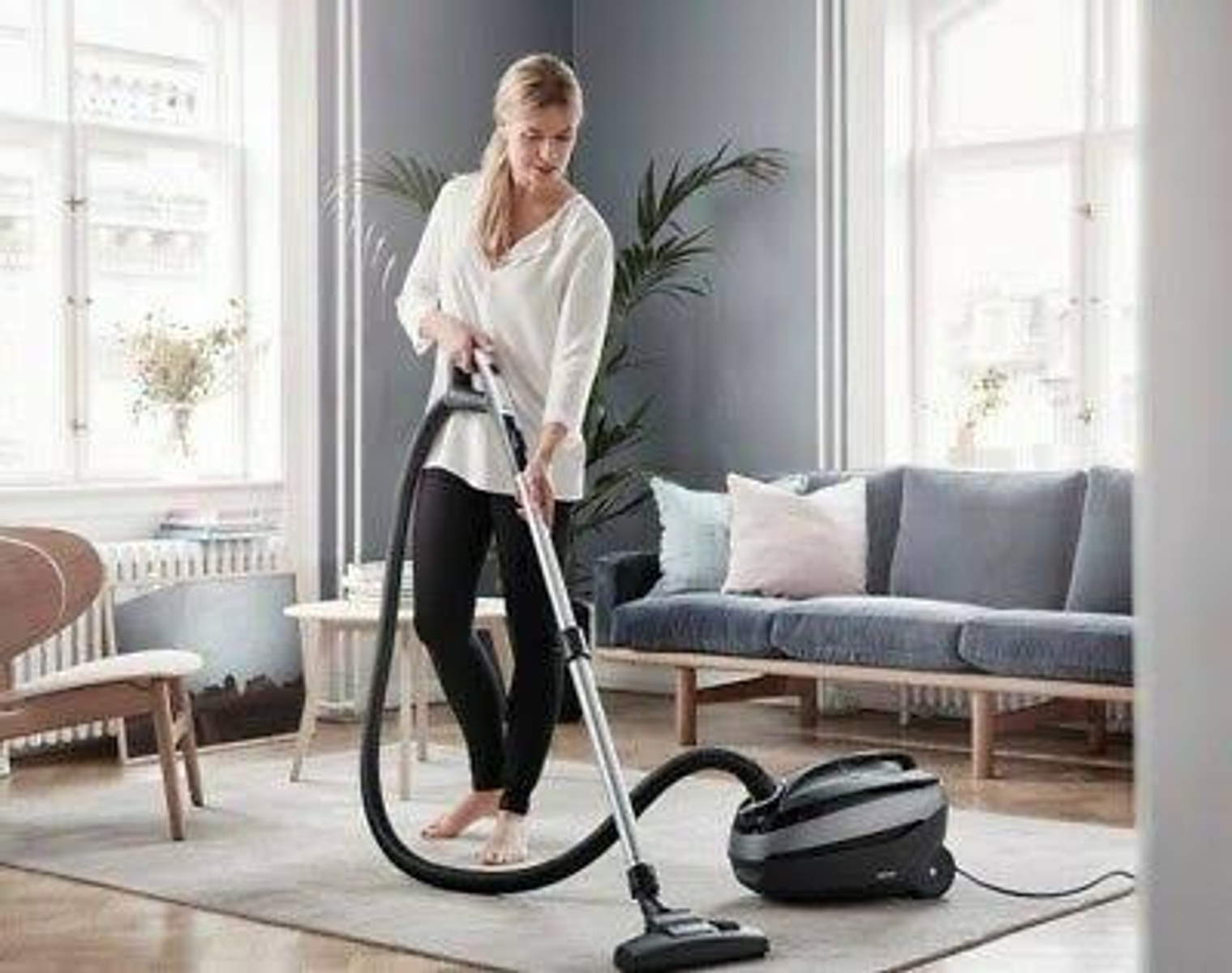 Una mujer aspirando una sala de estar alfombrada, sonriendo y sosteniendo la aspiradora.