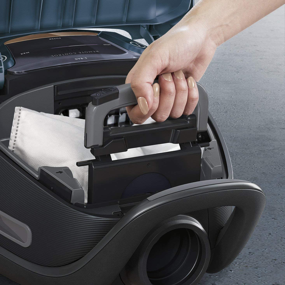 Ręka trzyma część odkurzacza, otwiera ją. Odkurzacz jest ciemnoszary, z białym filtrem w środku.