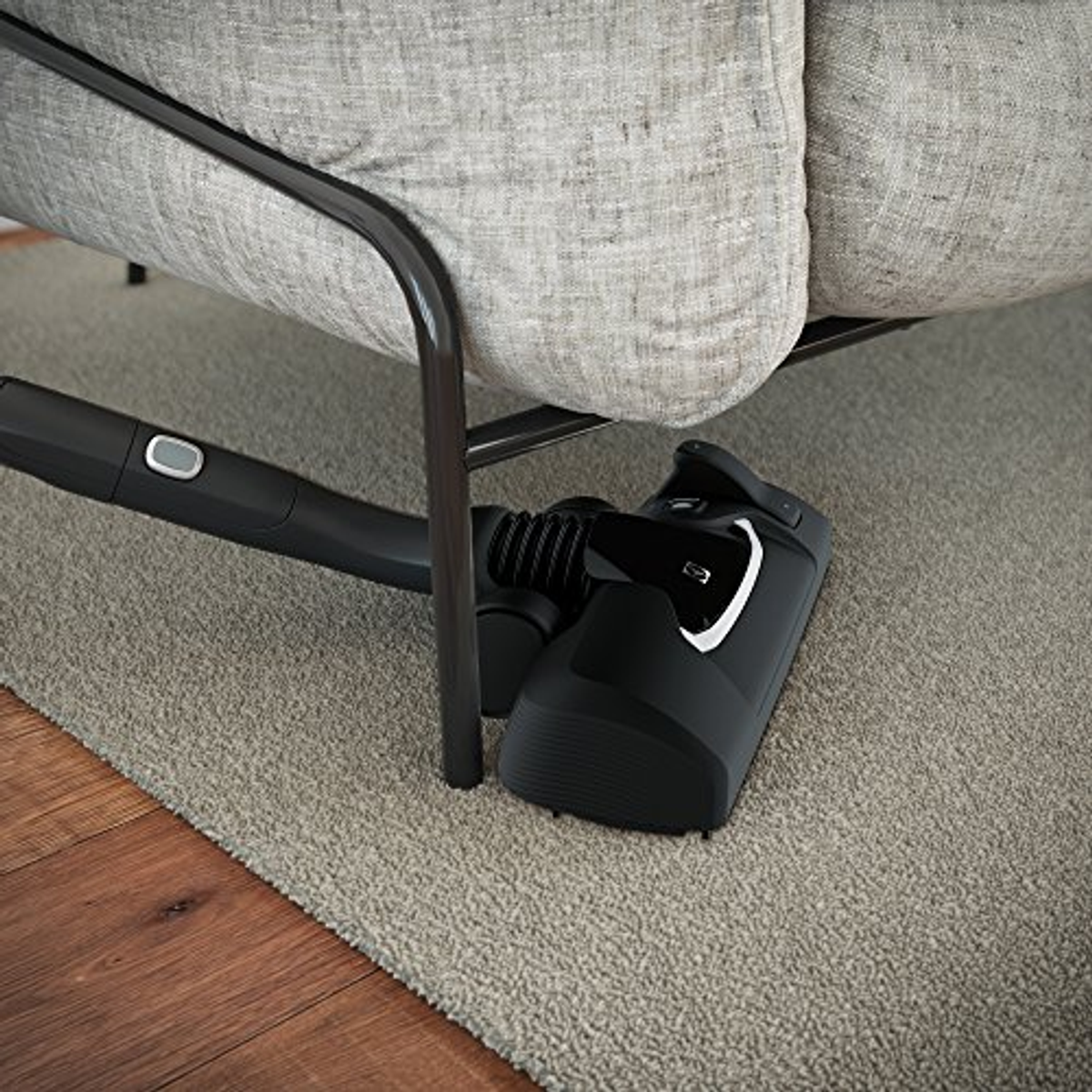 Czarna głowica odkurzacza czyszcząca szary dywan pod kanapą.