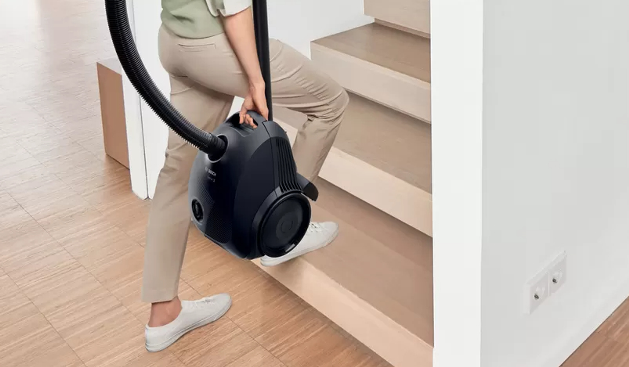 Osoba wnosząca czarny odkurzacz po drewnianych schodach.