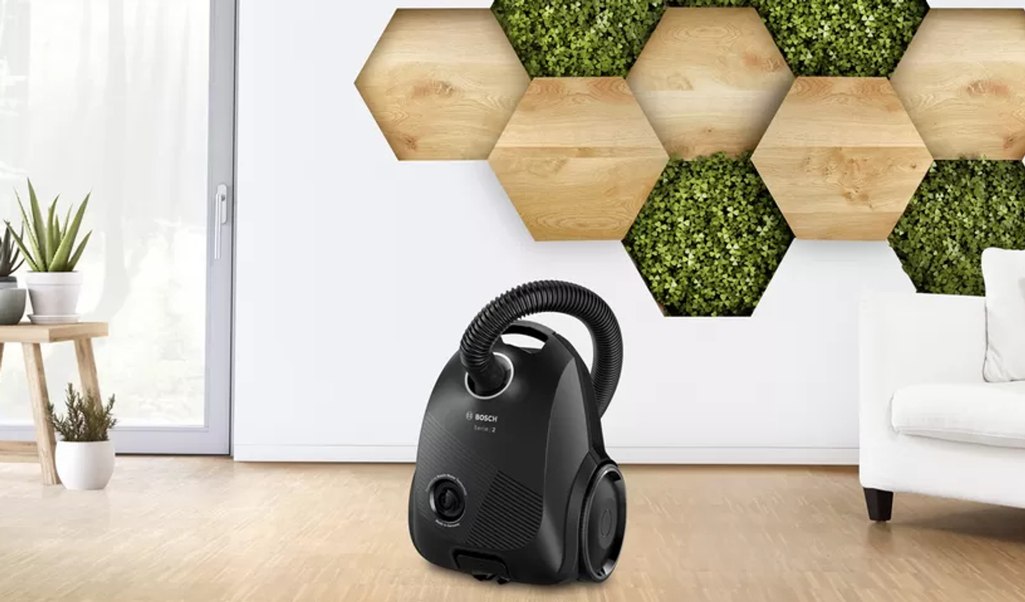 Czarny odkurzacz na drewnianej podłodze. Tło to biała ściana z wzorem plastra miodu i roślinami.