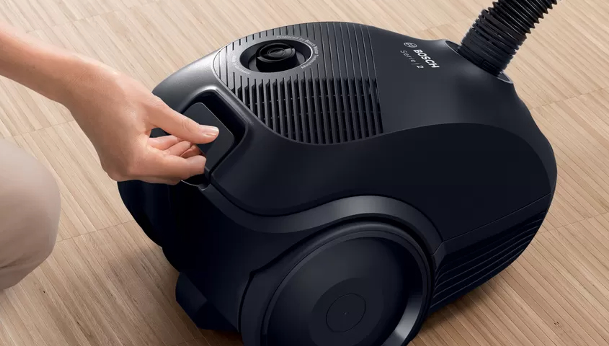 Czarny odkurzacz. Ręka otwiera pokrywę. Jest na drewnianej podłodze. Marka to Bosch.