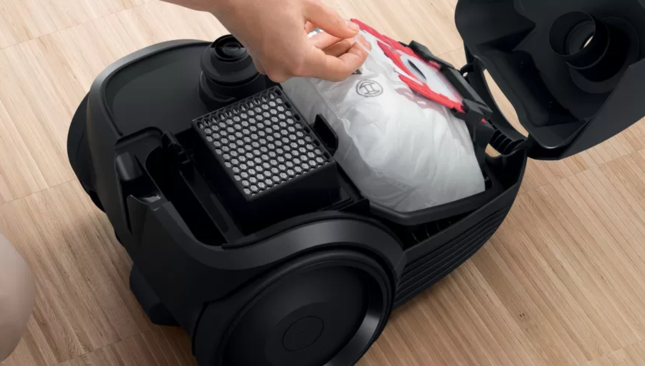 Czarny odkurzacz z otwartą komorą. Ręka trzyma białą torbę. Drewniana podłoga.