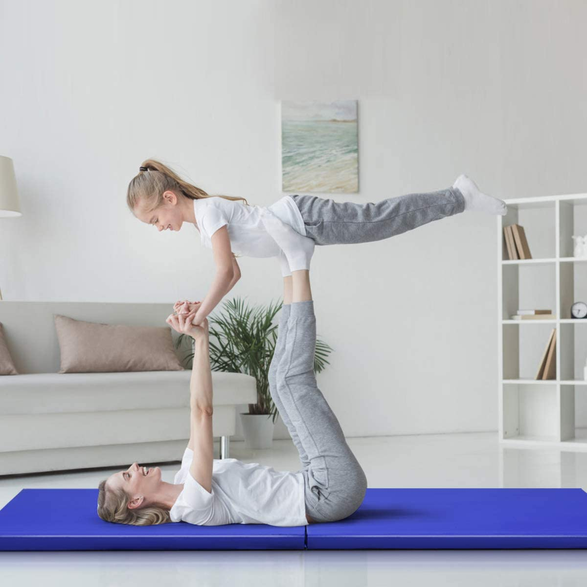 Frau auf einer blauen Matte, die ein Mädchen hochhebt. Sie machen eine Yoga-Pose in einem hellen Raum mit weißen Möbeln.