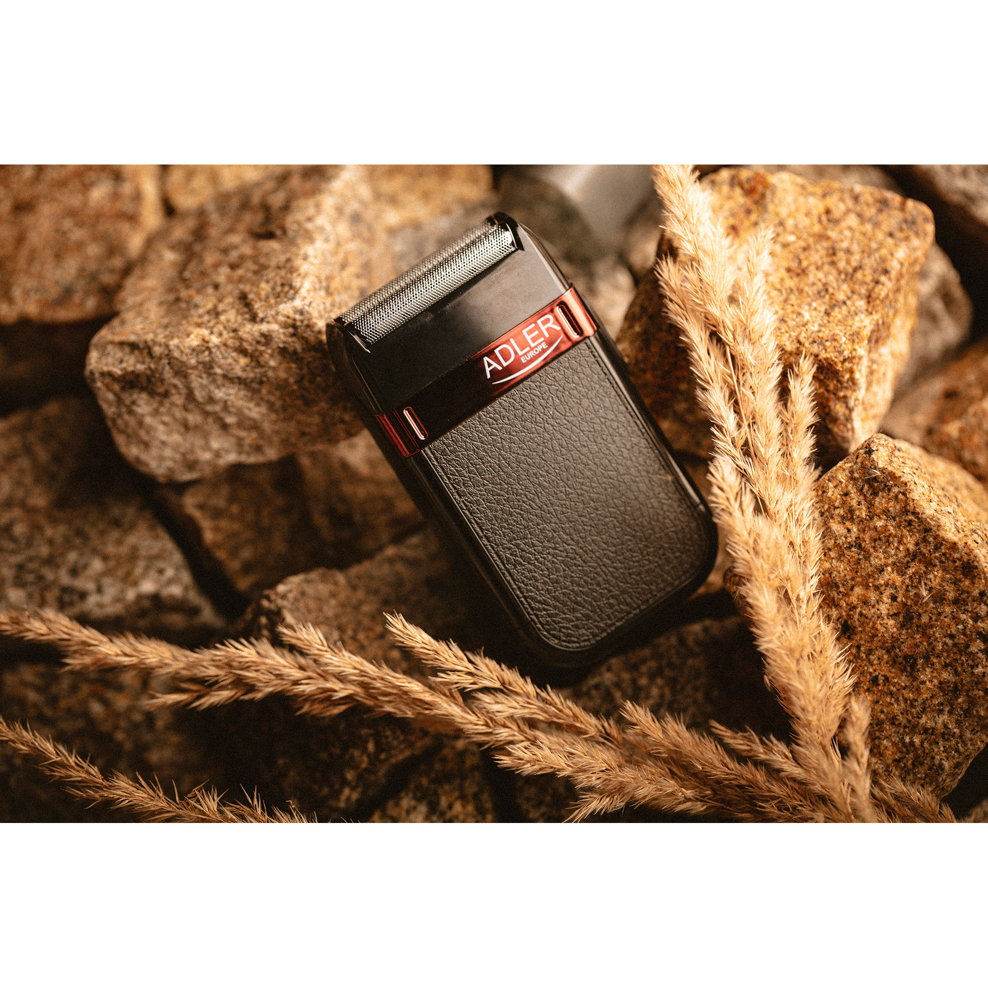 Czarna i czerwona golarka elektryczna ADLER na kamieniach z kilkoma suszonymi roślinami. Golarka ma czarny uchwyt.