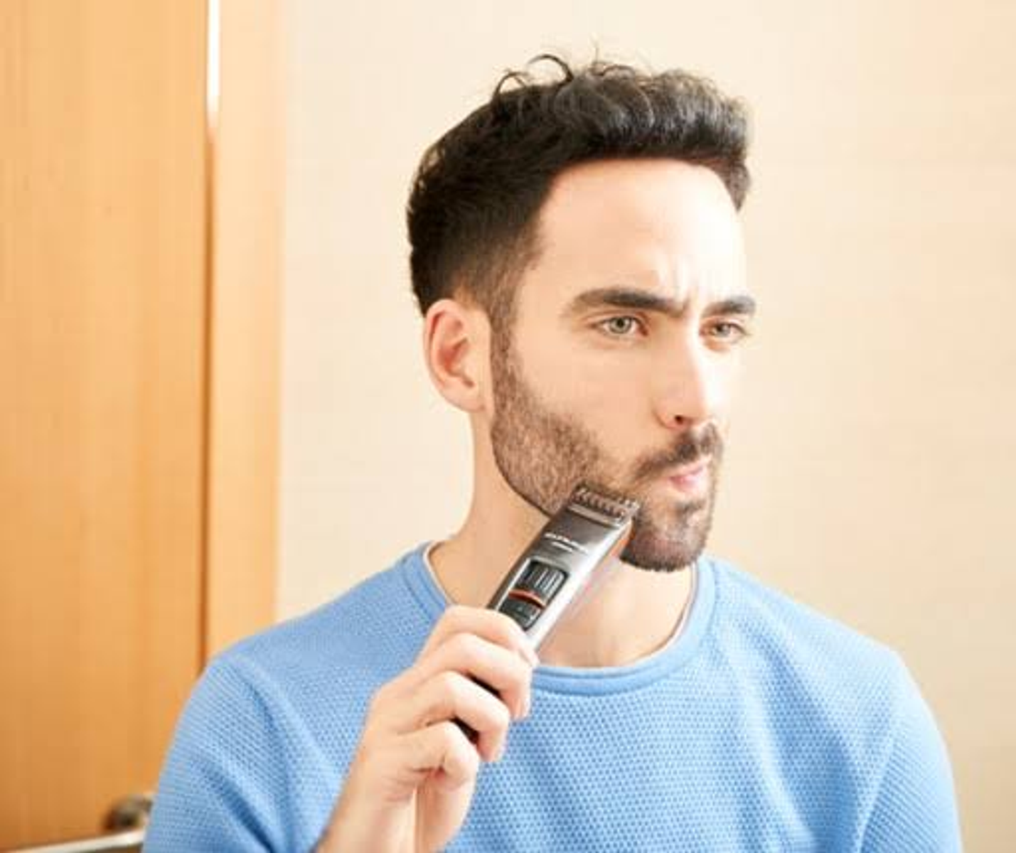 Un hombre se afeita la barba con una recortadora negra. Lleva un suéter azul. Fondo borroso.