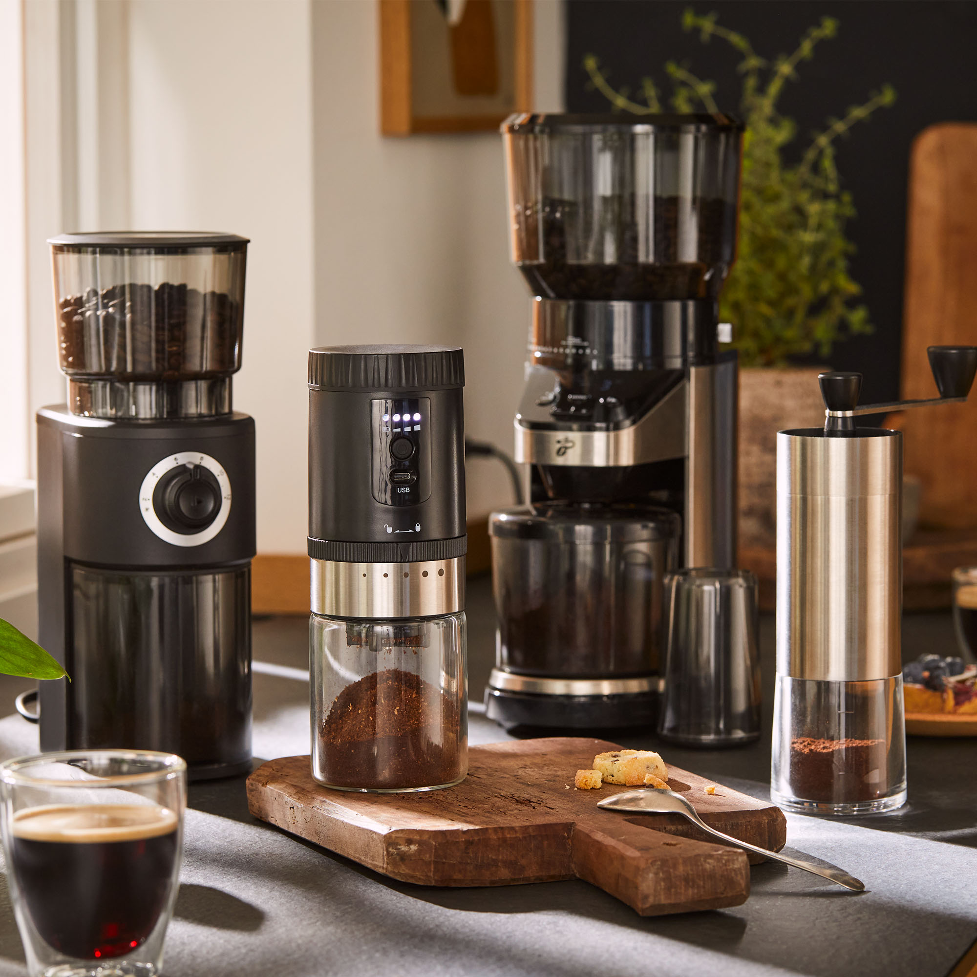 Kaffeemühlen und Kaffeemaschine auf einer Theke. Kaffeetasse und Gebäck im Vordergrund. Dunkler Hintergrund.