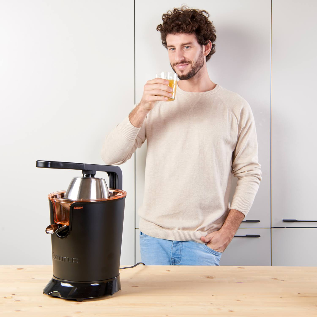 Un uomo beve succo da un bicchiere accanto a un estrattore di succo nero e arancione. Lo sfondo è una cucina.