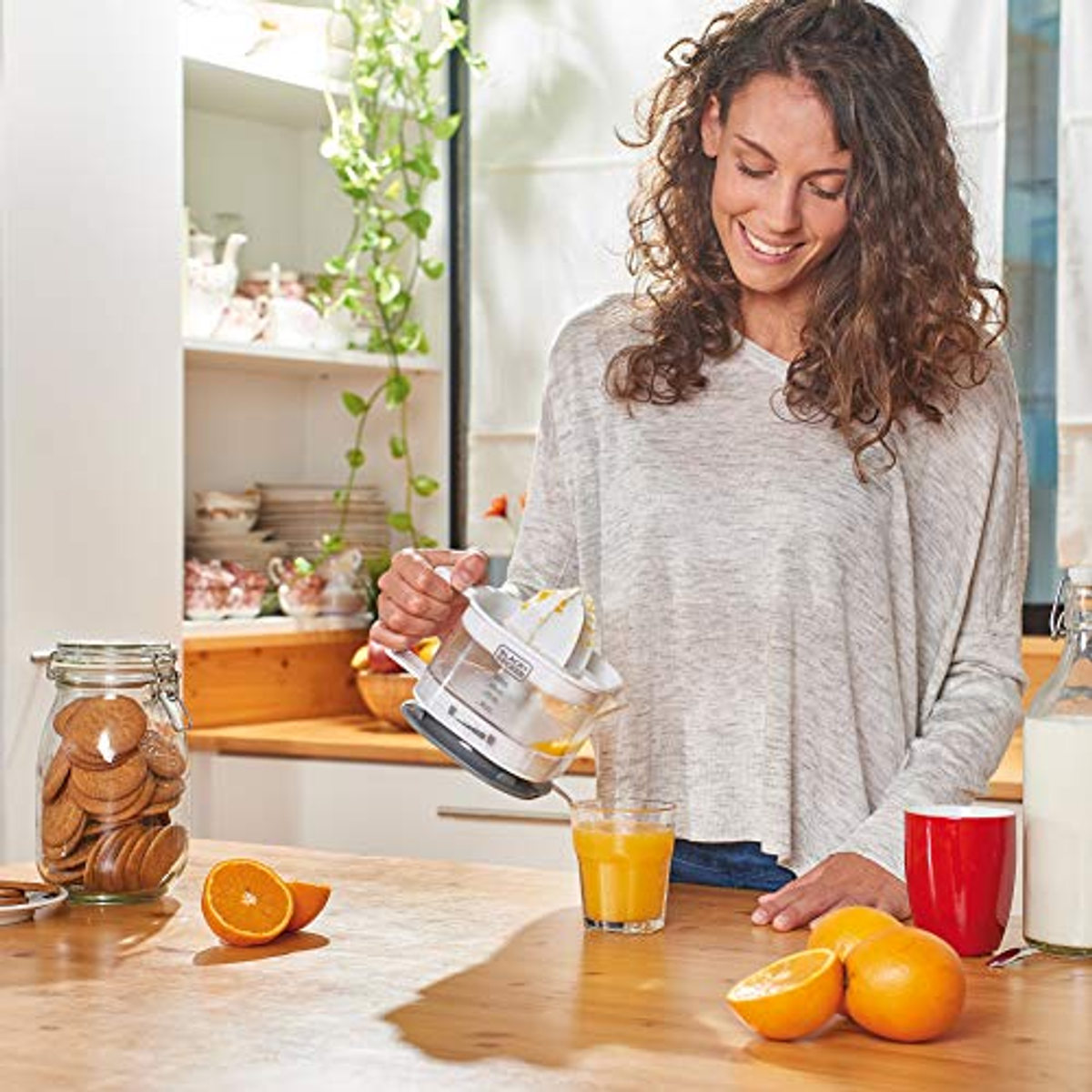 Woman juicing oranges in a kitchen. Includes oranges, juice, and a jar of cookies. Smiling.