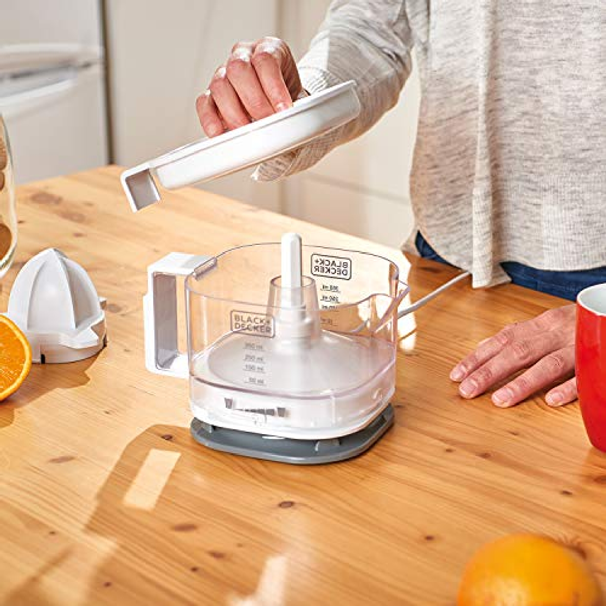 A woman adds a lid to a white food processor on a wooden table. An orange is nearby.