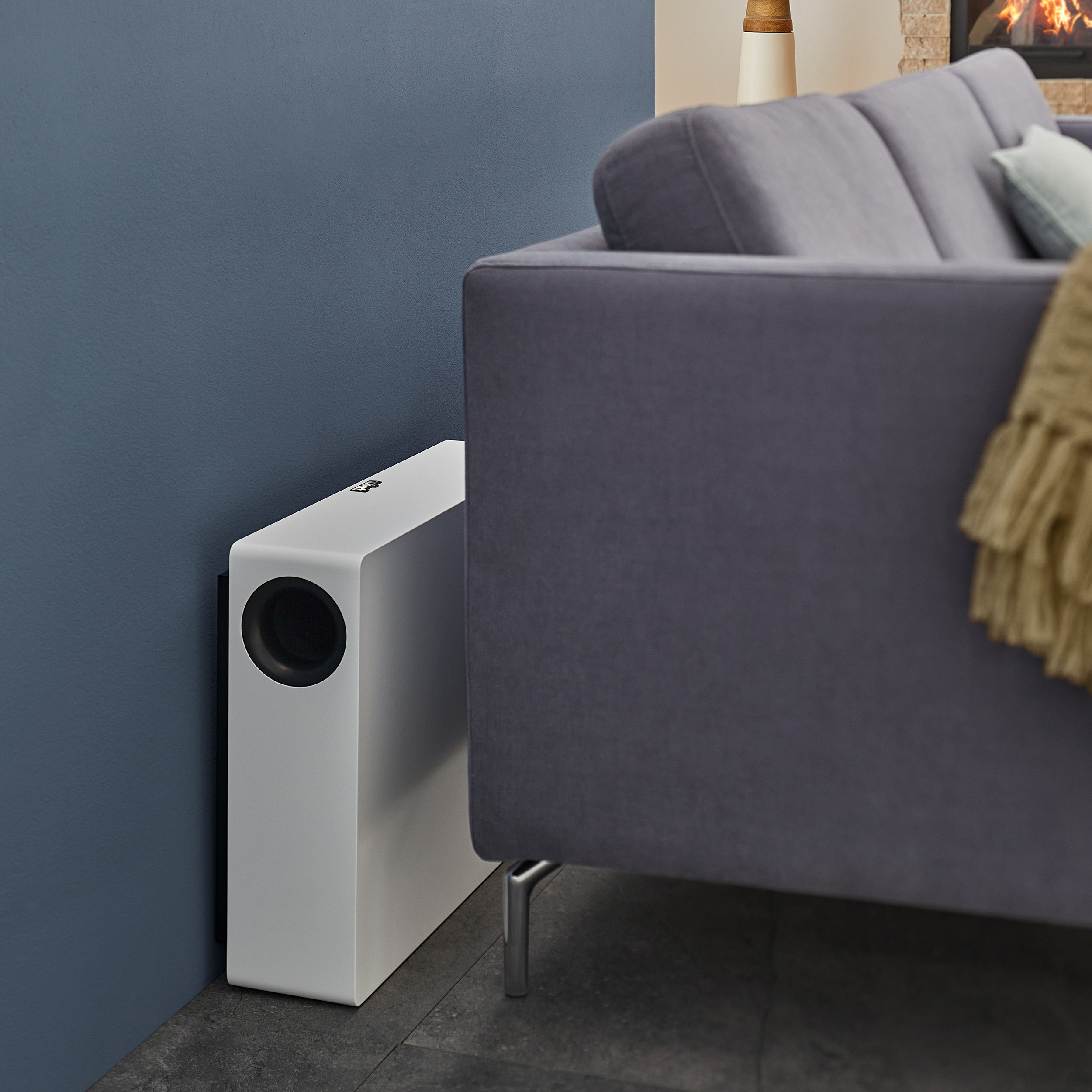 Ein weißer Subwoofer neben einem grauen Sofa und einer dunkelblauen Wand in einem Raum.