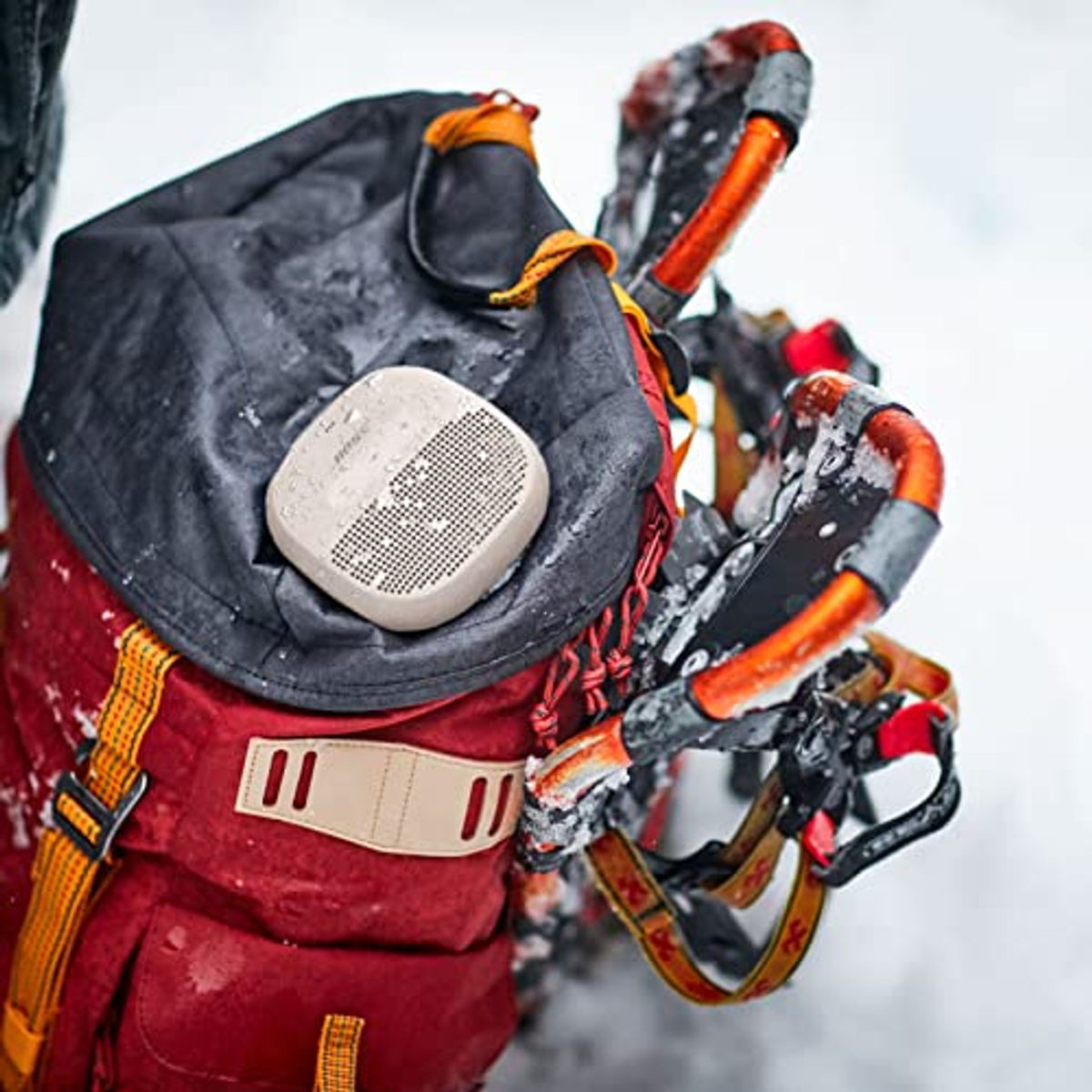Mały szary głośnik na czerwonym plecaku, ze śniegiem w tle. Scena na zewnątrz.