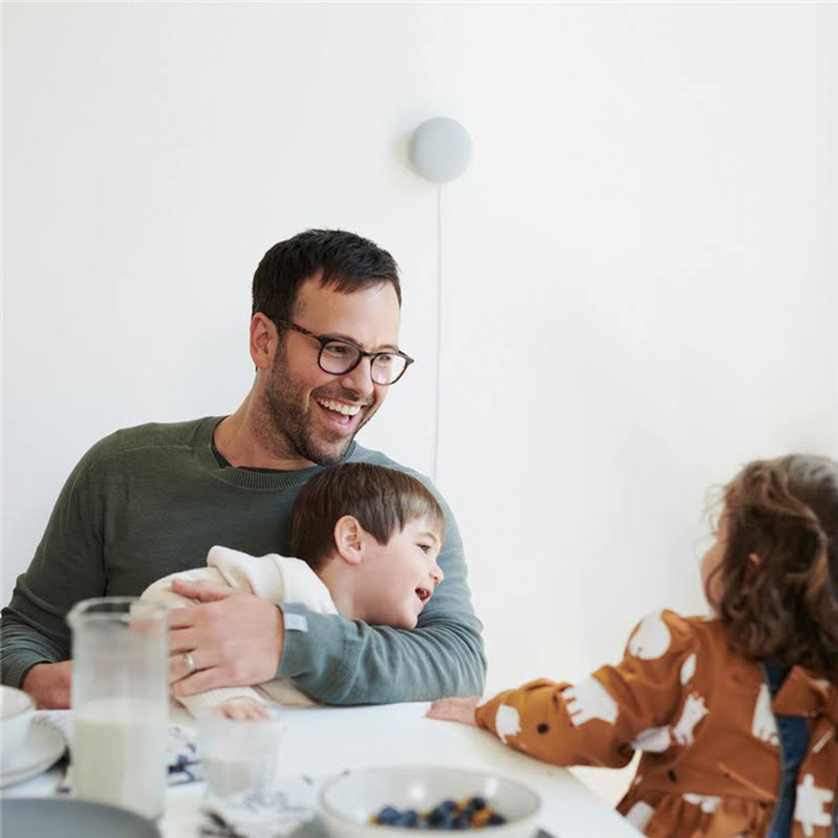 Ojciec obejmuje dwoje dzieci przy stole. Inteligentny głośnik jest zamontowany na ścianie.