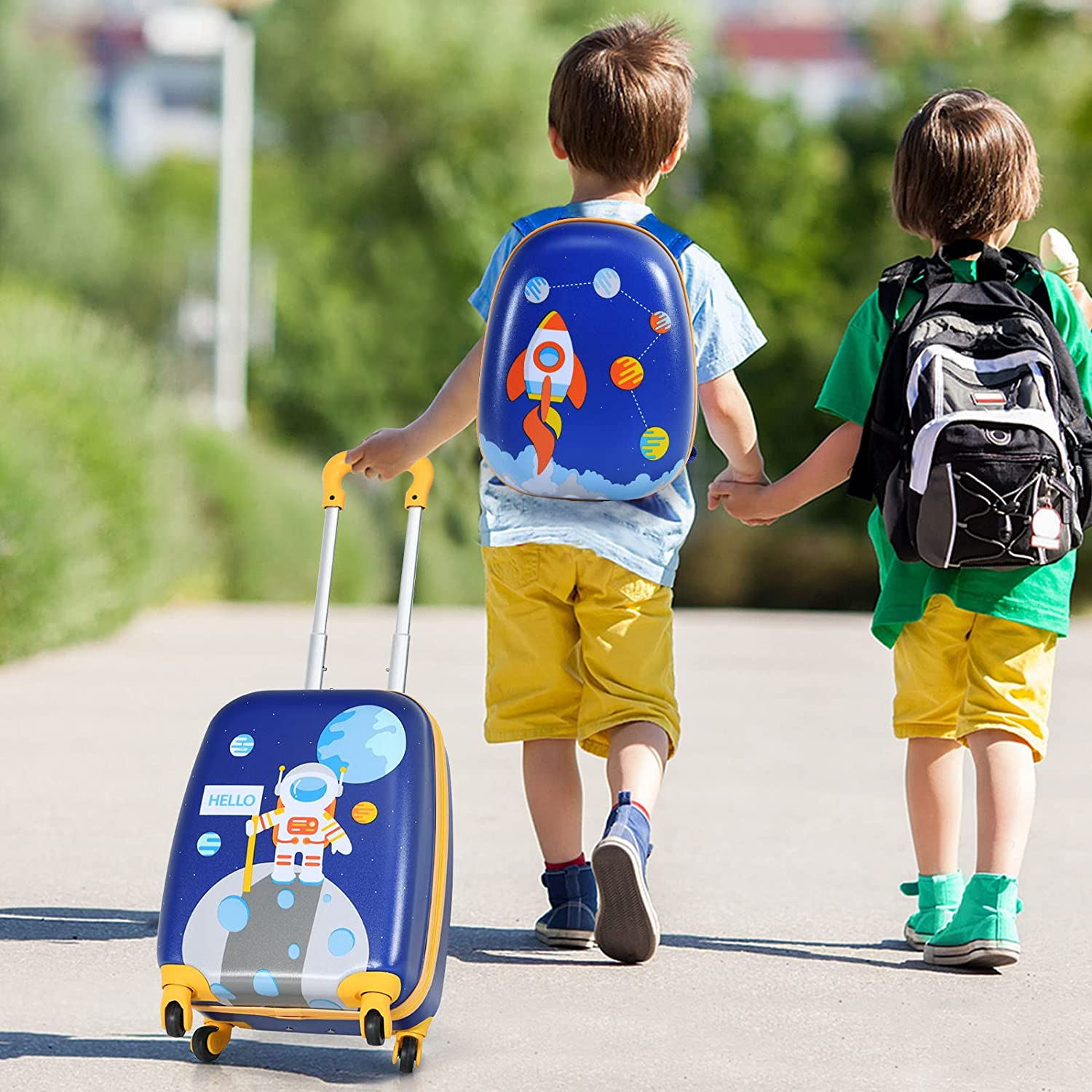 Zwei Kinder mit Rucksäcken gehen auf dem Bürgersteig, eines zieht einen blauen Koffer mit Astronautenkunst.