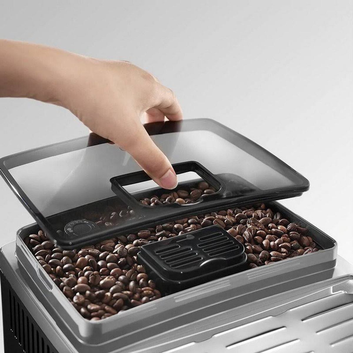 Una mano levanta la tapa transparente de un recipiente de granos de café, lleno de granos.