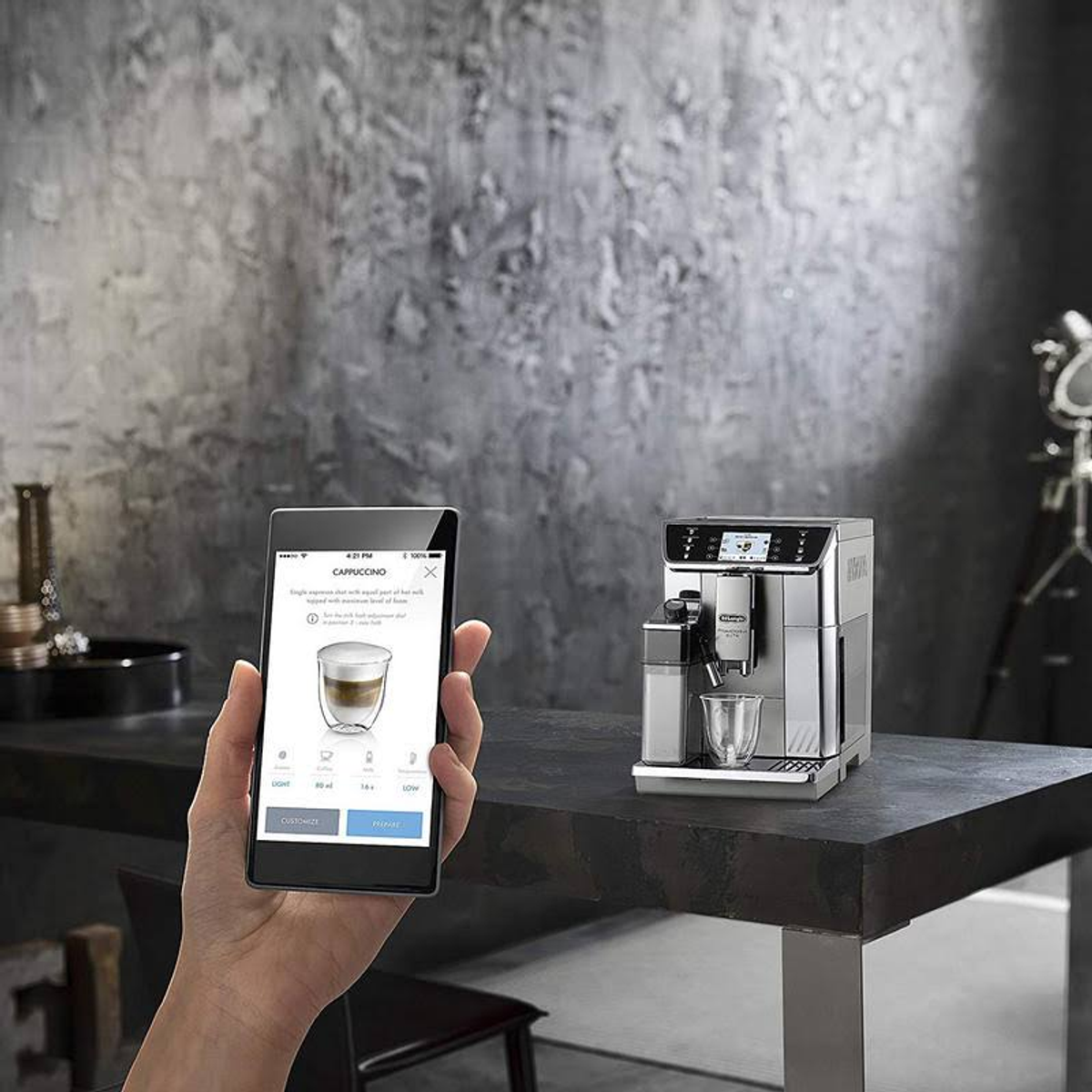 Eine Hand, die ein Smartphone hält und eine Cappuccino-Maschine steuert.