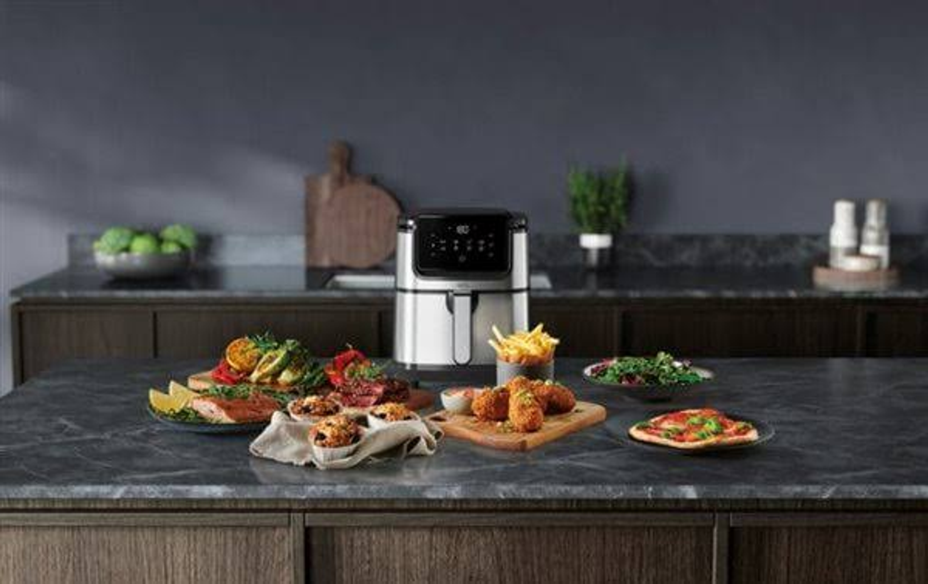 Una friggitrice ad aria con vari cibi, tra cui verdure e pizza, disposti su un bancone.