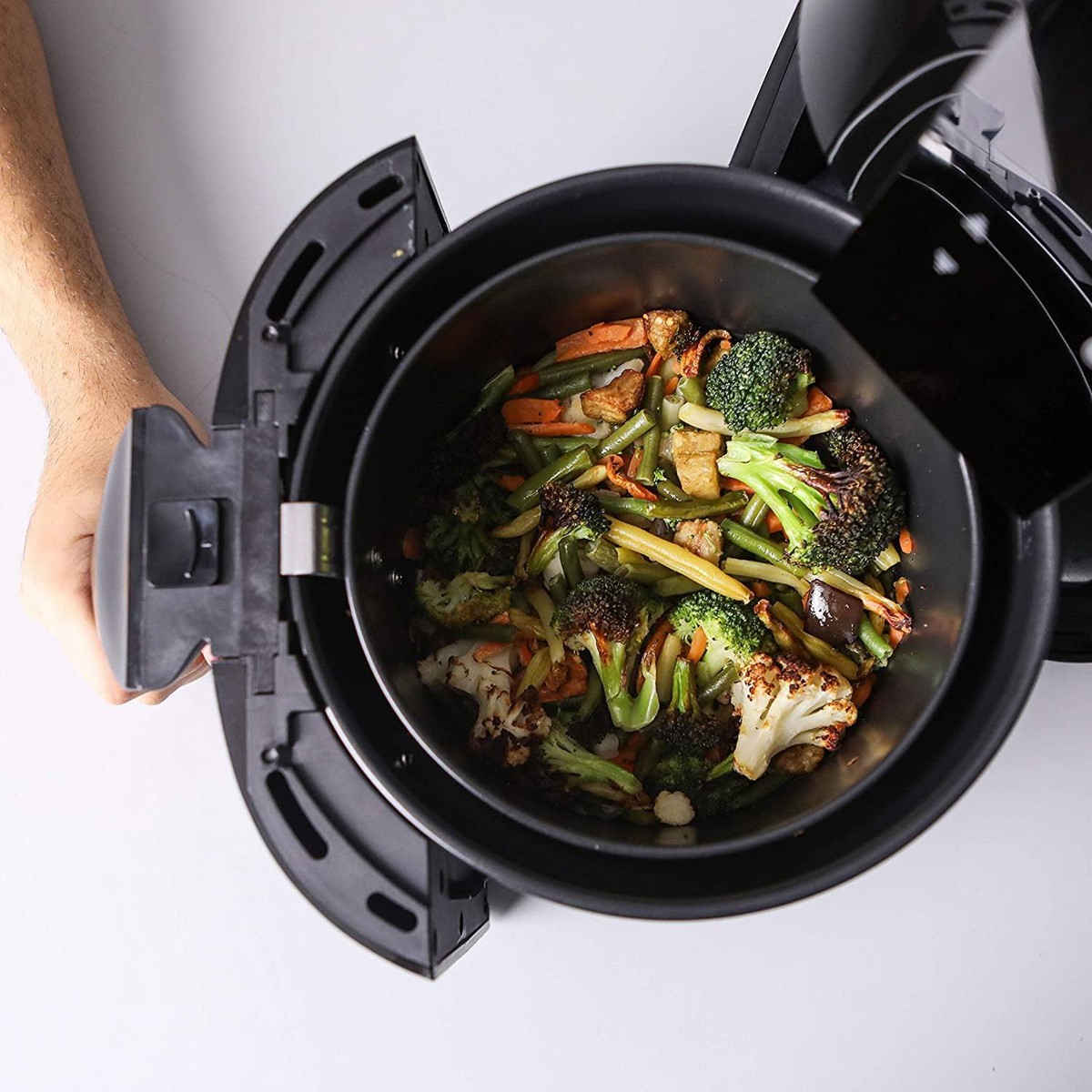 Una cesta de freidora abierta con verduras cocinadas. Una mano sostiene la freidora.