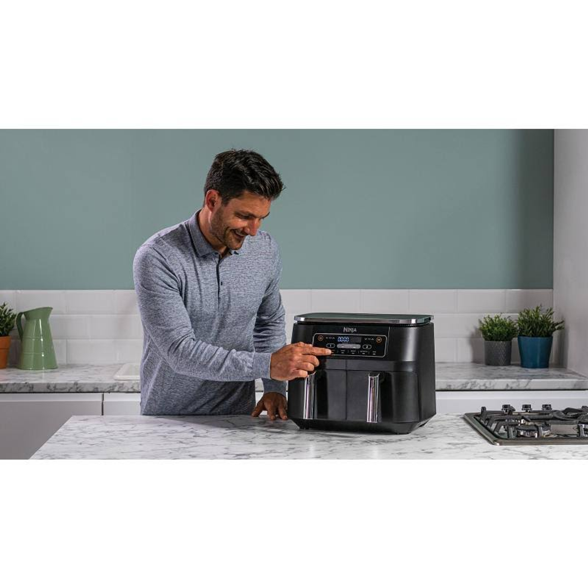 Mann benutzt schwarzen Airfryer auf weißer Arbeitsplatte. Pflanzen, Herd im Hintergrund.