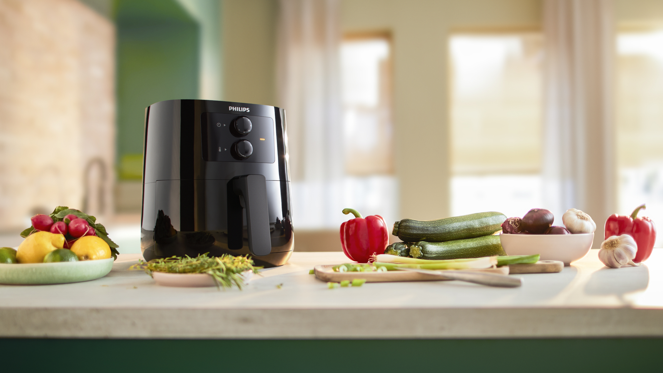 Freidora de aire negra con verduras e ingredientes en mostrador blanco, con fondo soleado.