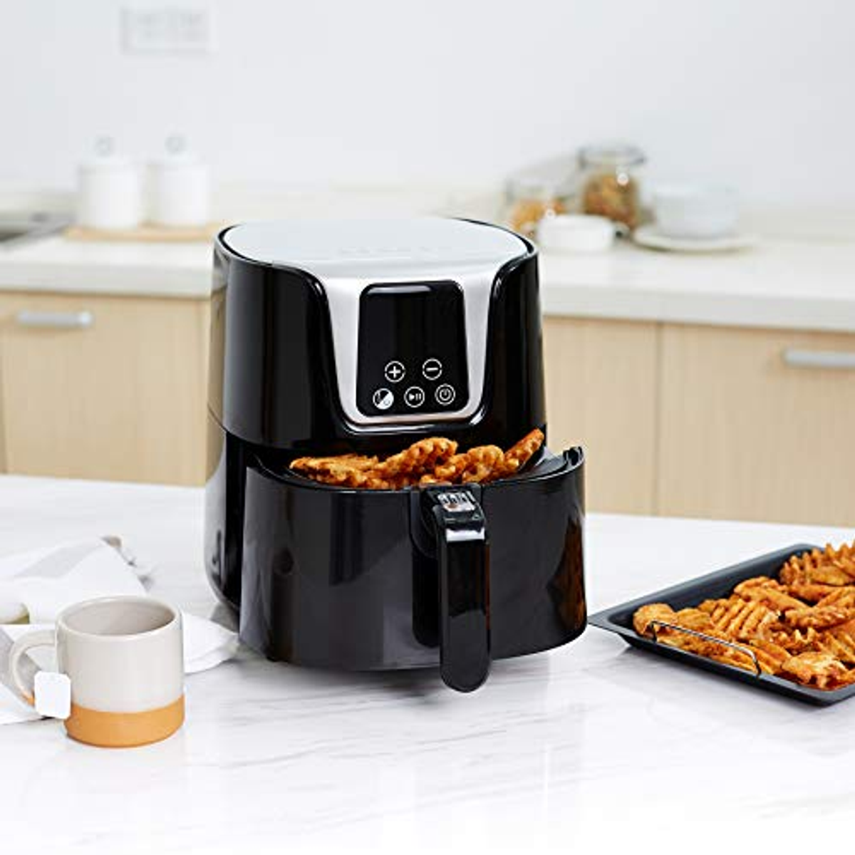 Friggitrice ad aria nera con patatine fritte. Una tazza bianca e un vassoio di patatine fritte sono sul tavolo. Sfondo della cucina luminoso.