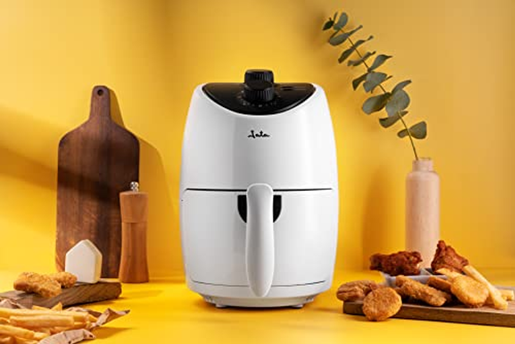 Weißer Airfryer mit schwarzen Akzenten, gebratene Speisen vorne. Gelber Hintergrund mit Schneidebrett und Pflanze.