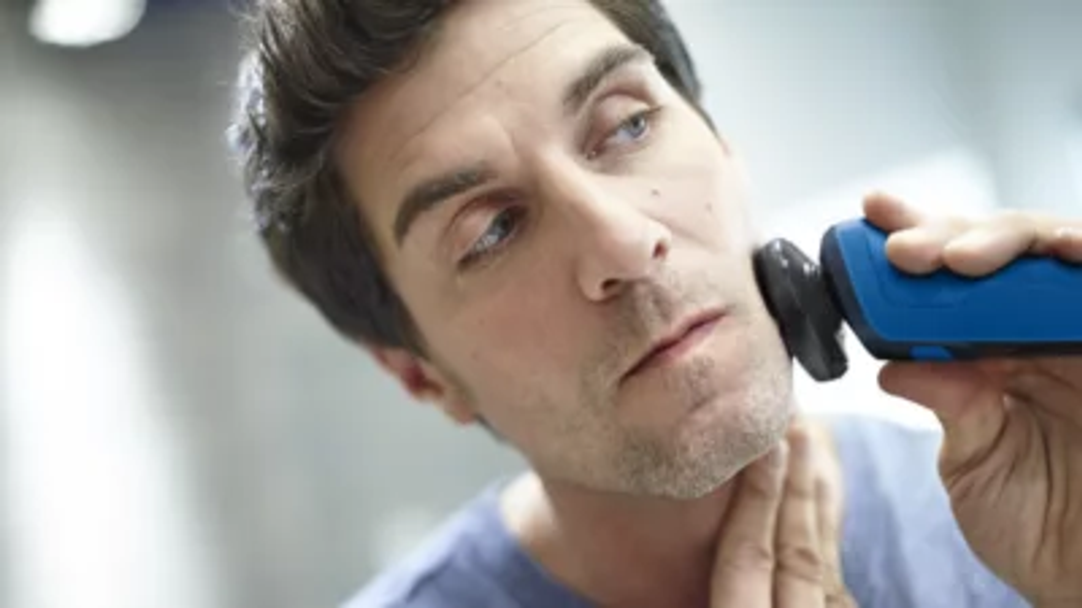 Mężczyzna goli brodę niebieską golarką elektryczną. Ujęcie z bliska. Wygląda poważnie.