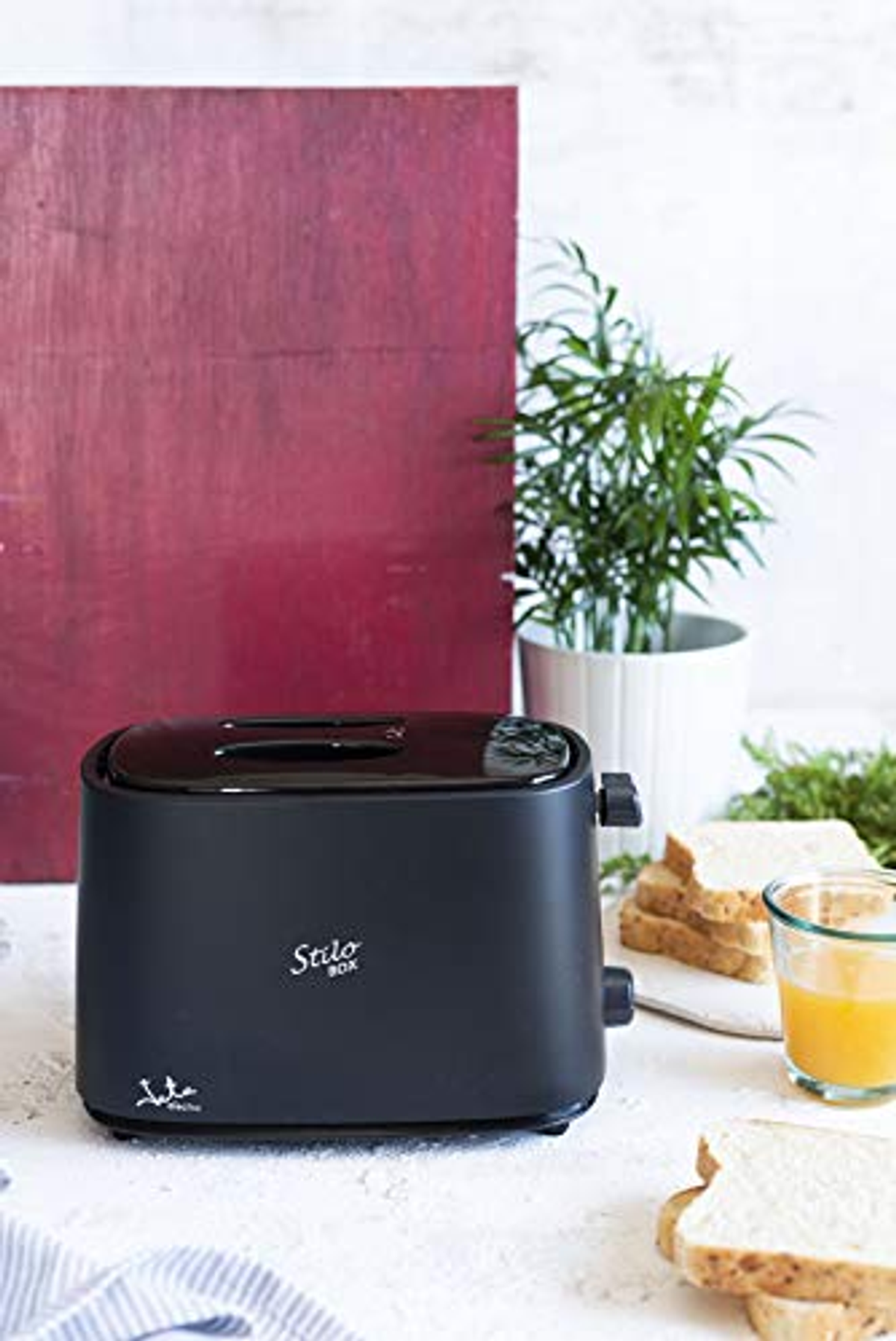 Czarny toster na białej powierzchni z tostami, sokiem pomarańczowym i rośliną. Czerwone tło.