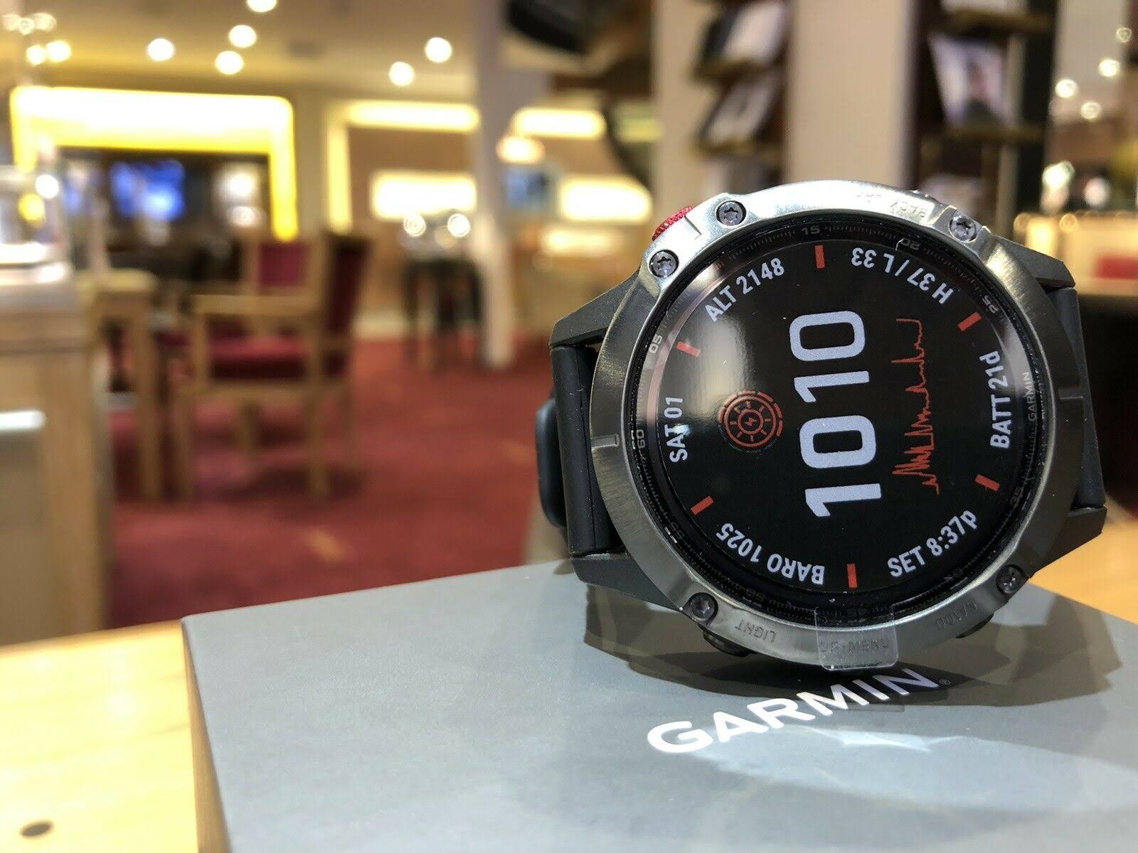 Garmin-Smartwatch auf einer Box, die Daten anzeigt. Der Hintergrund zeigt ein verschwommenes Interieur mit Lichtern und Tischen.