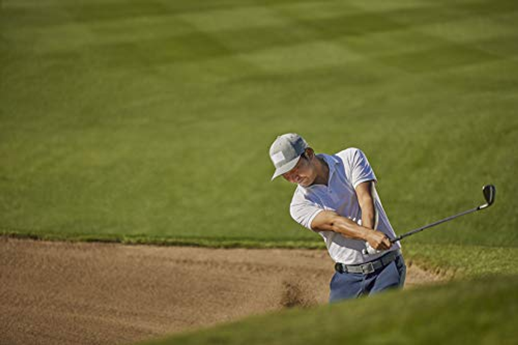 Un golfista en una trampa de arena, balanceando un palo en un campo verde.